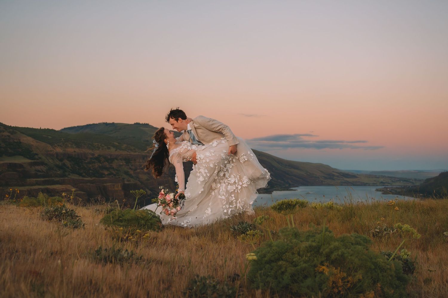 Couple sharing a romantic kiss at sunset on a hilltop with scenic water views on their wedding day.