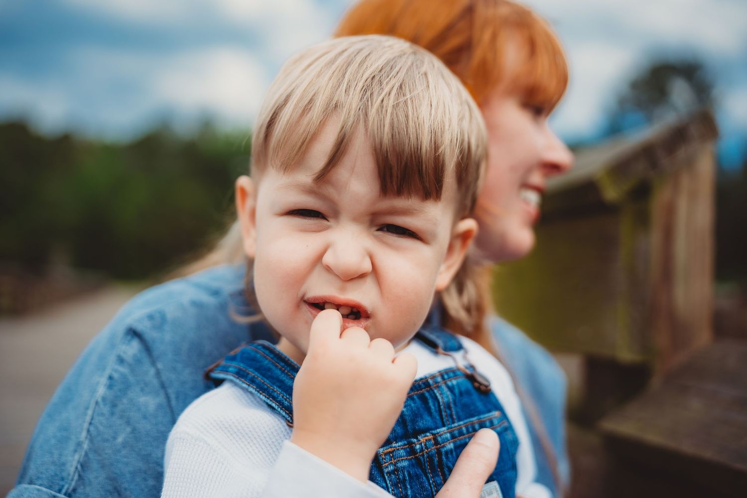 A toddler in denim overalls biting their fingers with a scrunched face, held by a redheaded woman outdoors.
