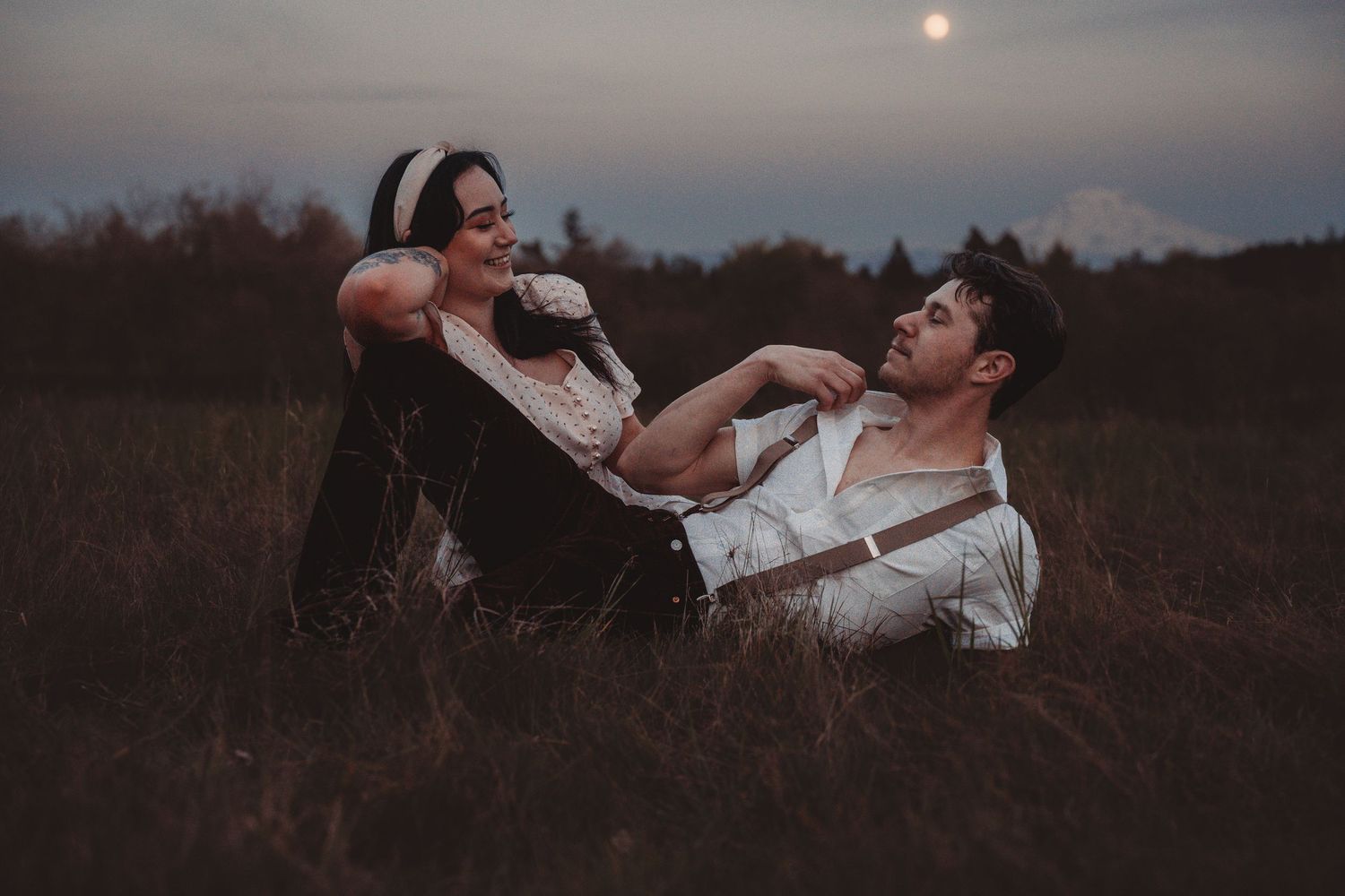 Couple playfully laughing together while sitting in a field at twilight with moon in background.