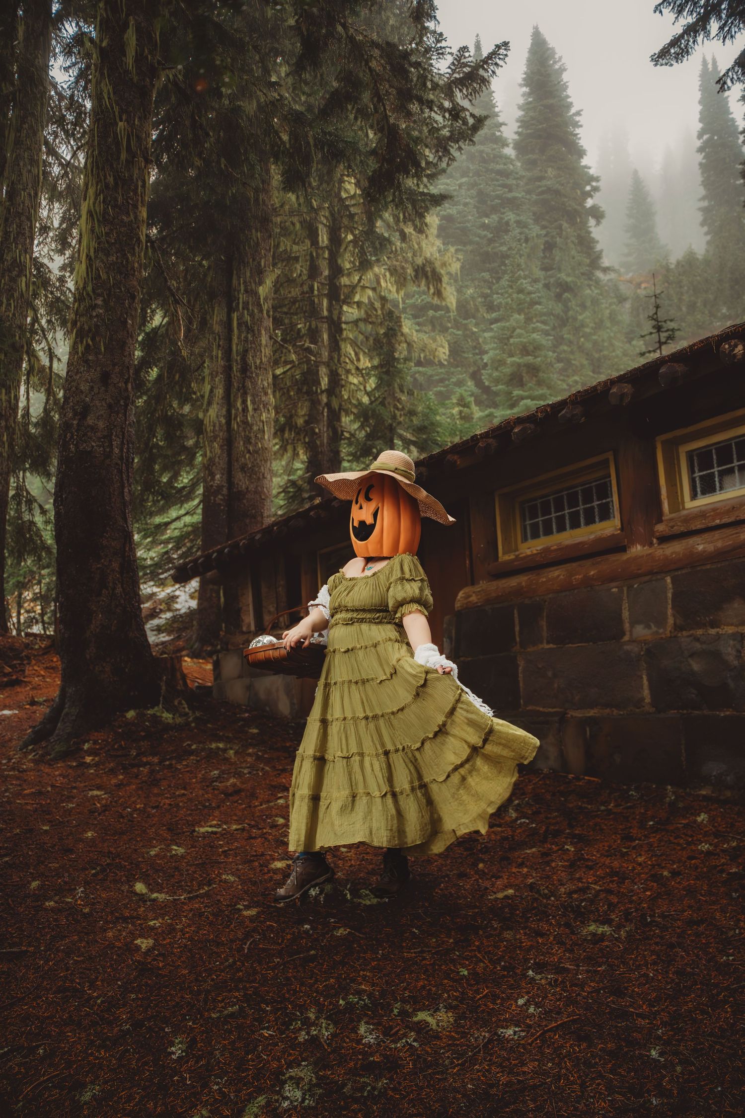 Woman wearing pumpkin head costume in green dress and hat standing near rustic cabin in foggy forest. mount Rainer national park