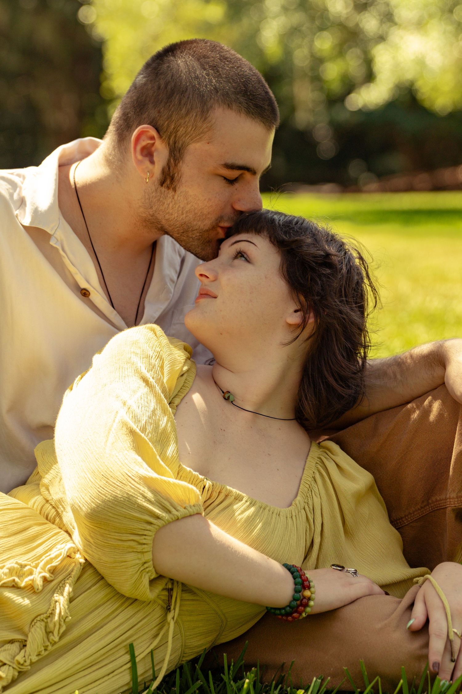 A couple shares a tender kiss while relaxing on green grass in a sunny park, woman in yellow dress.