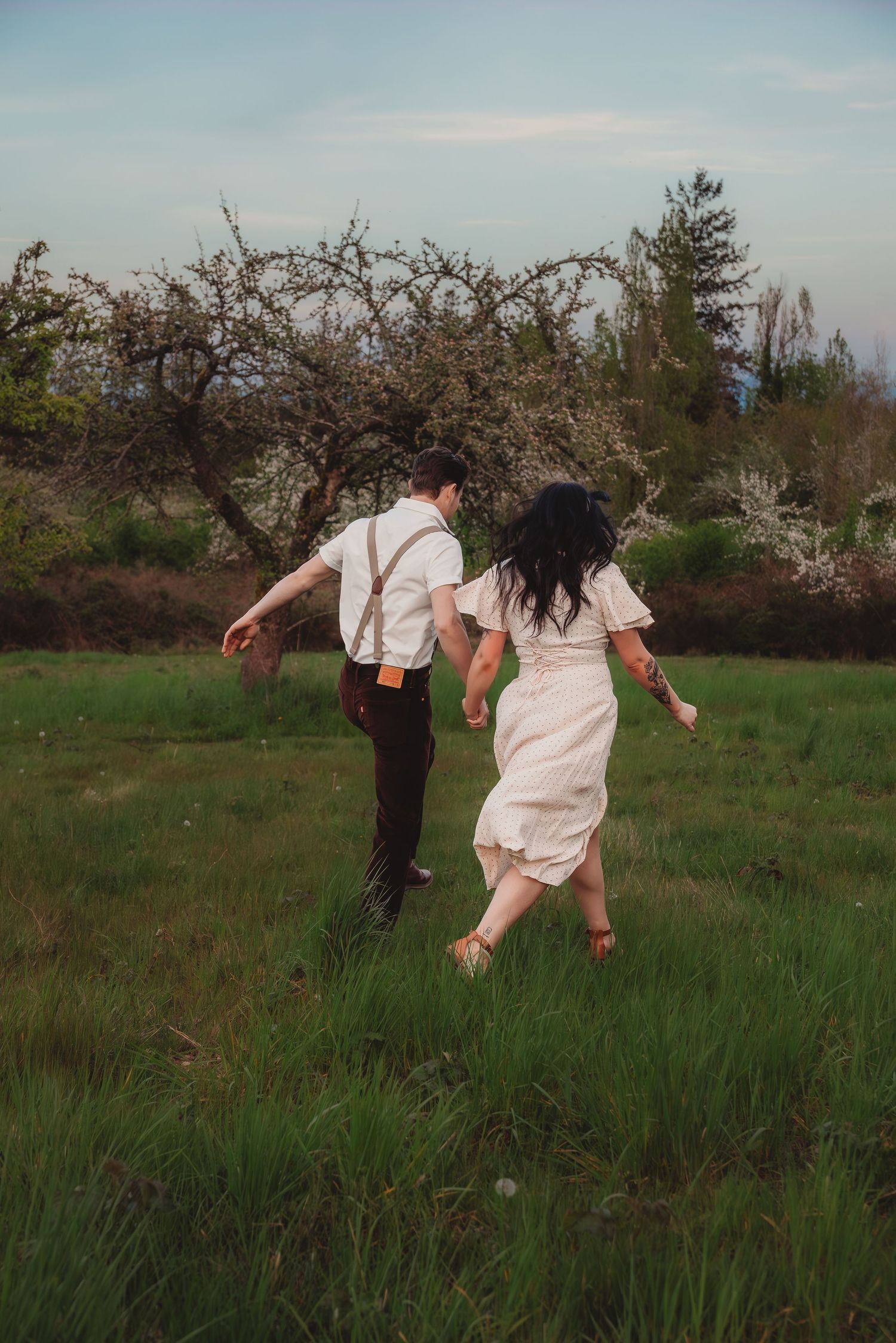 Couple running hand-in-hand through a green meadow with blooming trees at dusk.