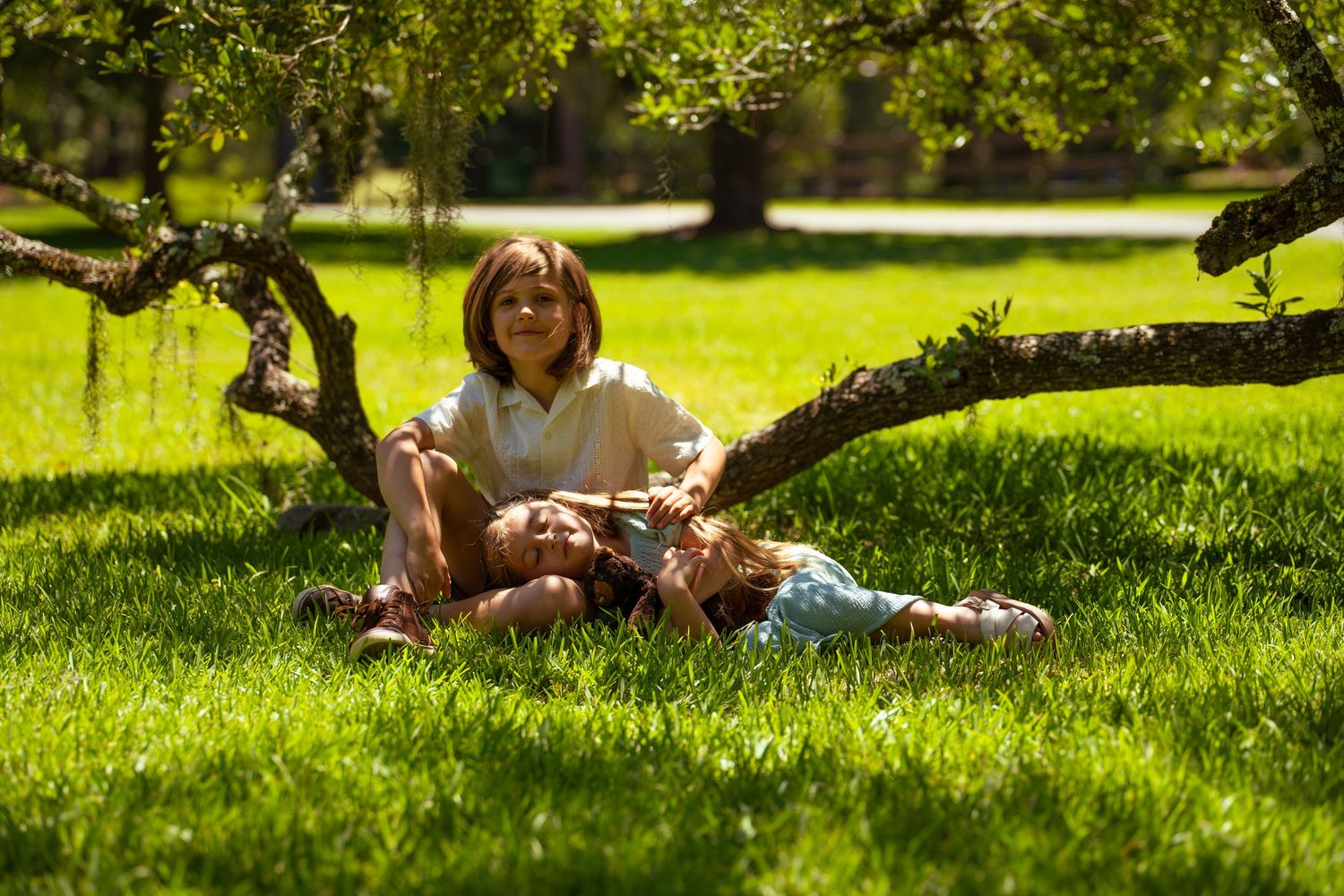 Two children relaxing under a tree on lush green grass in a sunny park setting.