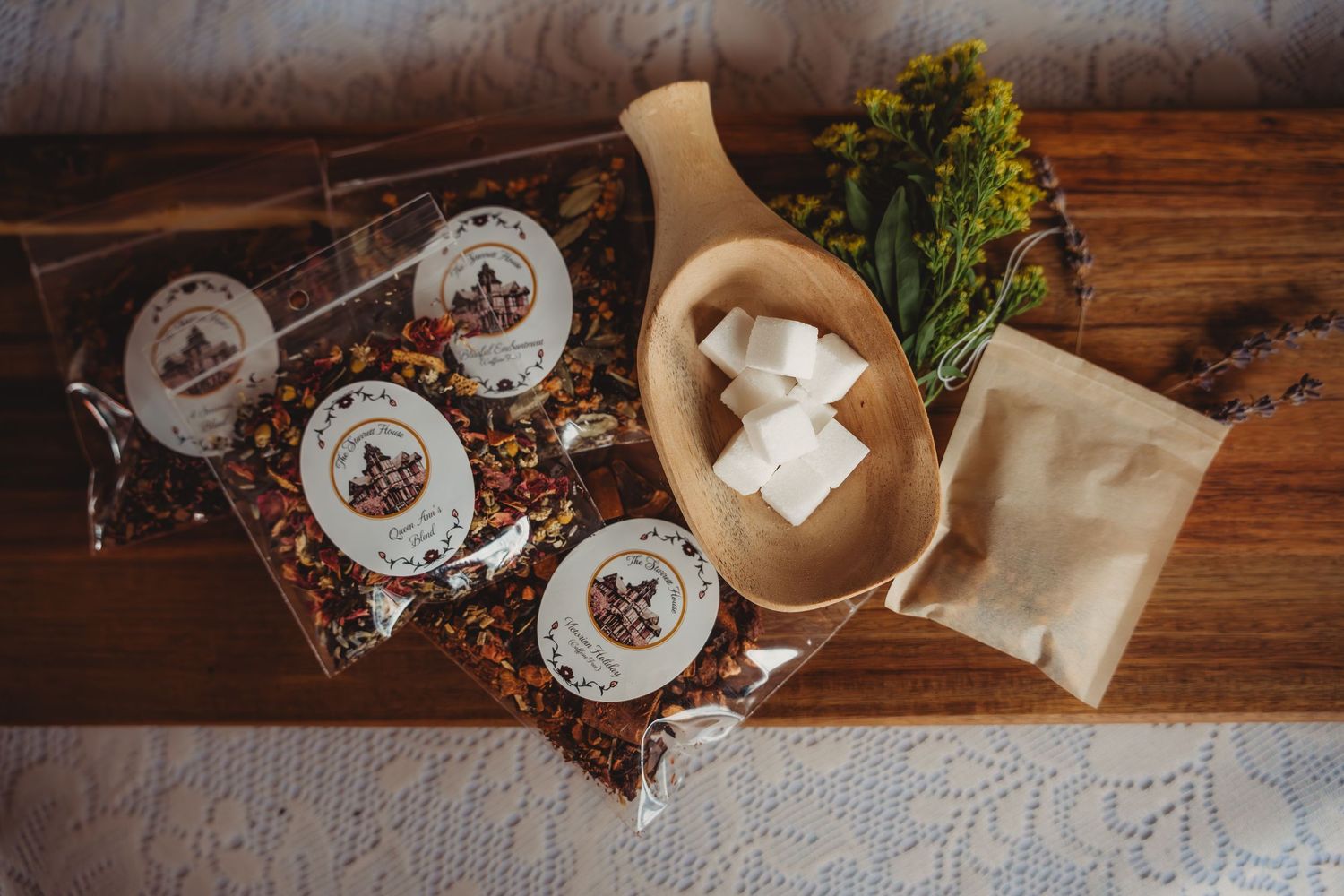 Artisan tea gift set with labeled spice packets, wooden bowl with sugar cubes, dried flowers, and kraft bag on wooden tray.