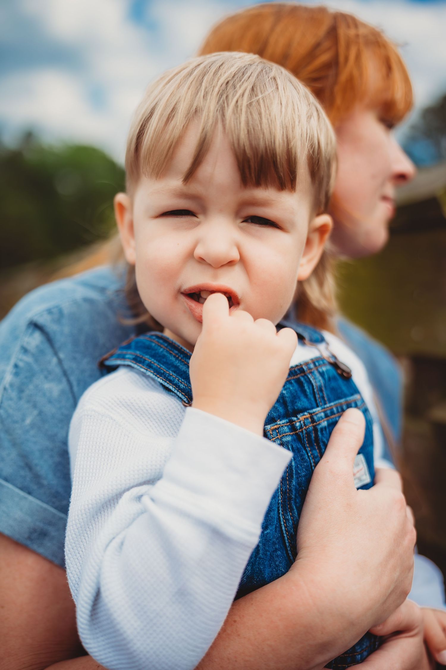 Toddler in denim overalls with finger in mouth being held by redheaded parent outdoors.