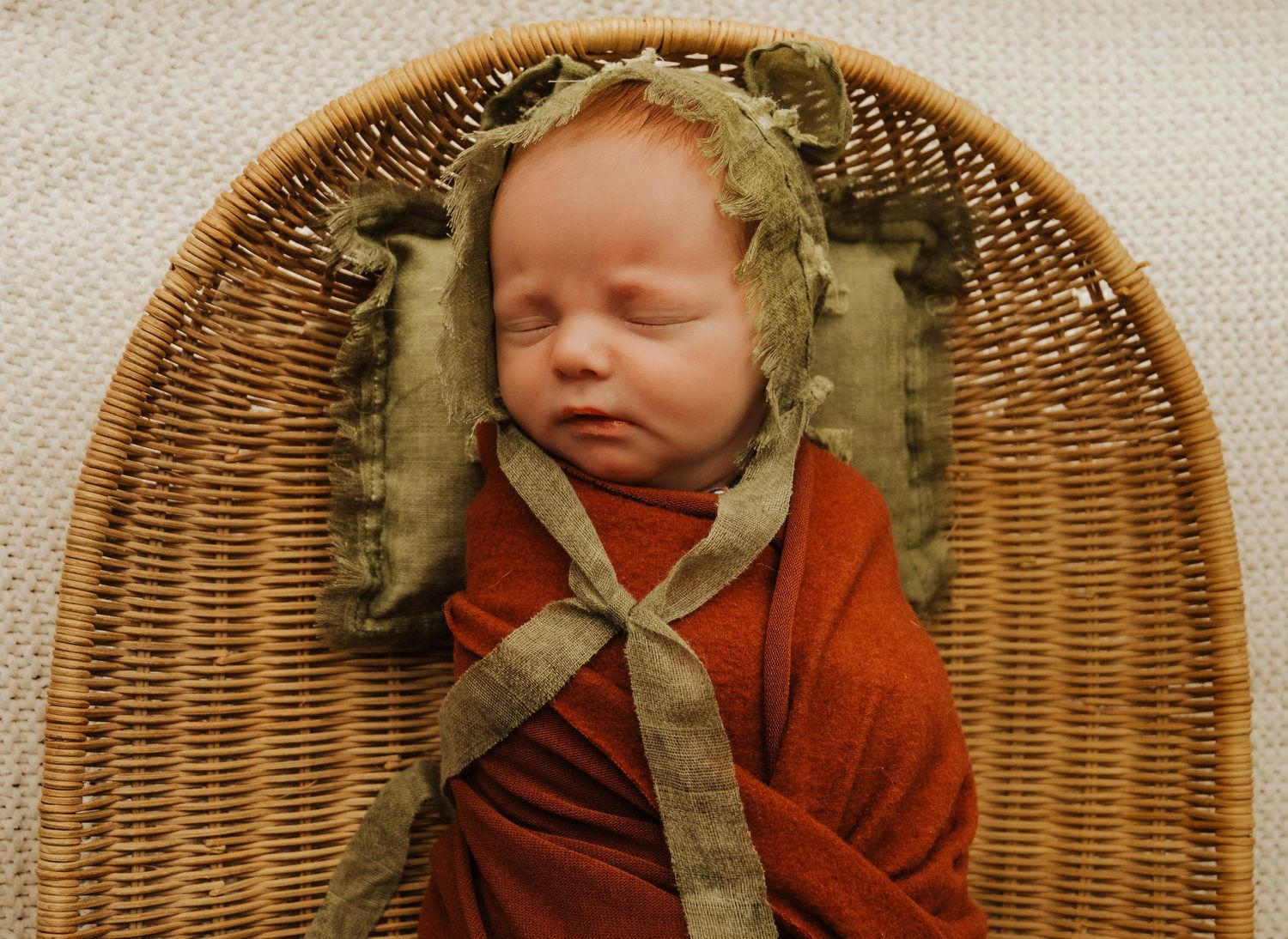 Newborn baby sleeping peacefully in a wicker basket, wrapped in a rust swaddle with a green bonnet.
