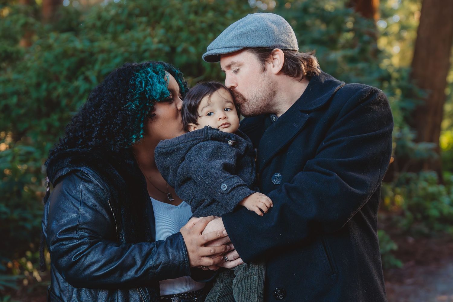 A couple sharing a tender moment kissing their baby outdoors, surrounded by lush green trees.