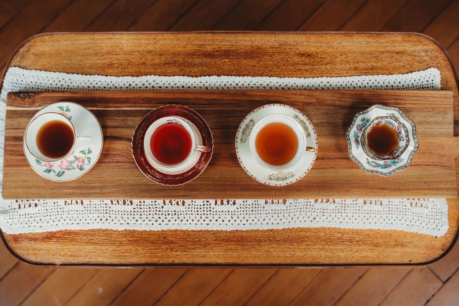 Four vintage teacups filled with dark tea arranged in a row on a wooden board with lace runner.