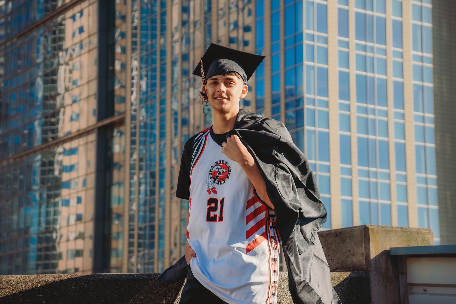 Young man in graduation cap and sports jersey number 21 posing confidently in front of city skyscrapers.