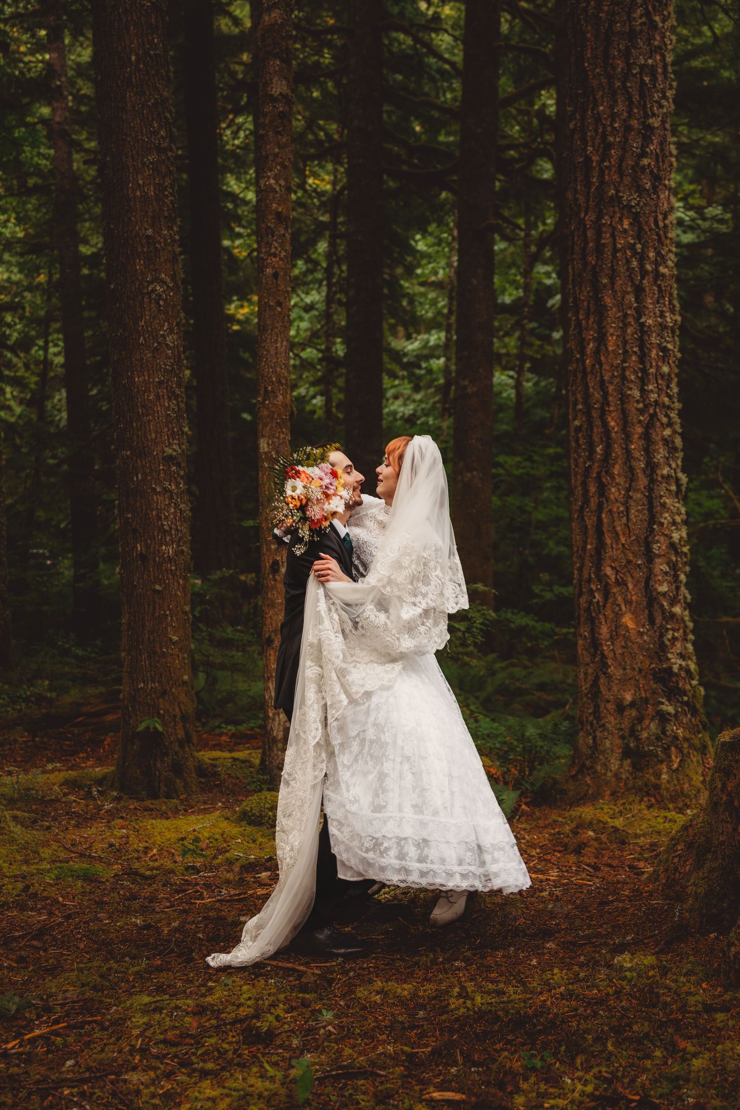A bride and groom share a romantic embrace in a lush forest, the bride wearing a lace gown and veil, holding a floral bouquet.