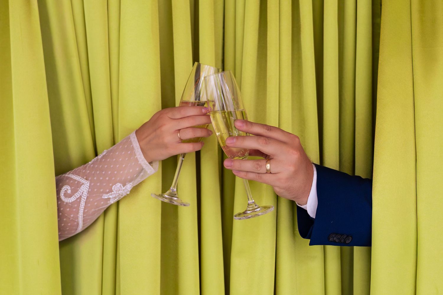 Two hands clinking champagne glasses in a celebratory toast against a yellow-green curtain backdrop.