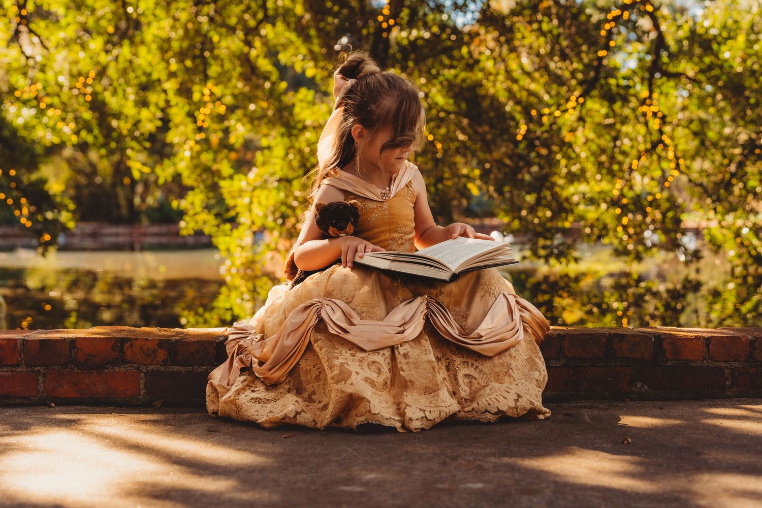 A young girl in a golden princess gown sits on a brick ledge reading a book outdoors near a sunlit pond.