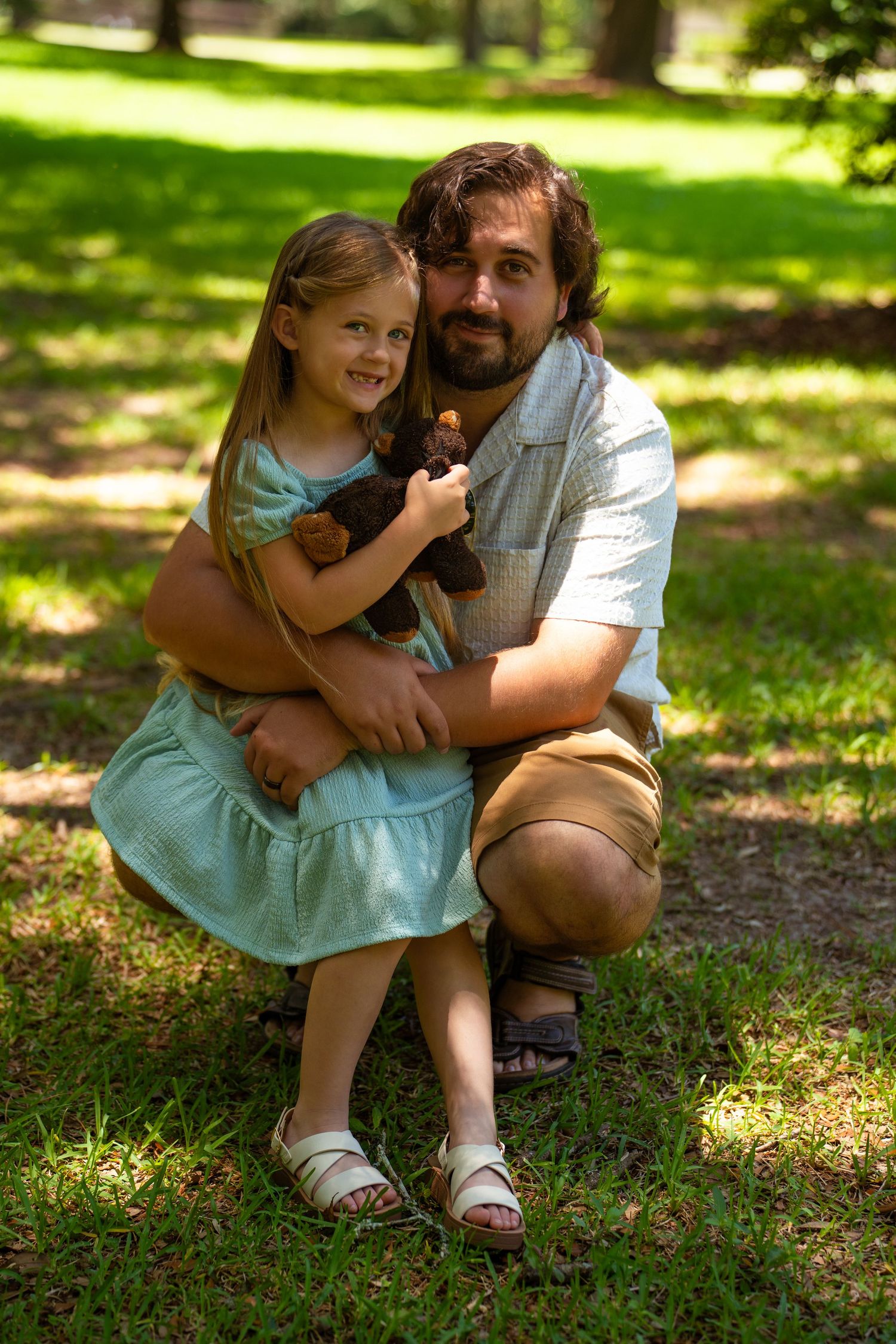 A father crouches beside his young daughter in a sunny park, hugging her as she holds a teddy bear in a teal dress.