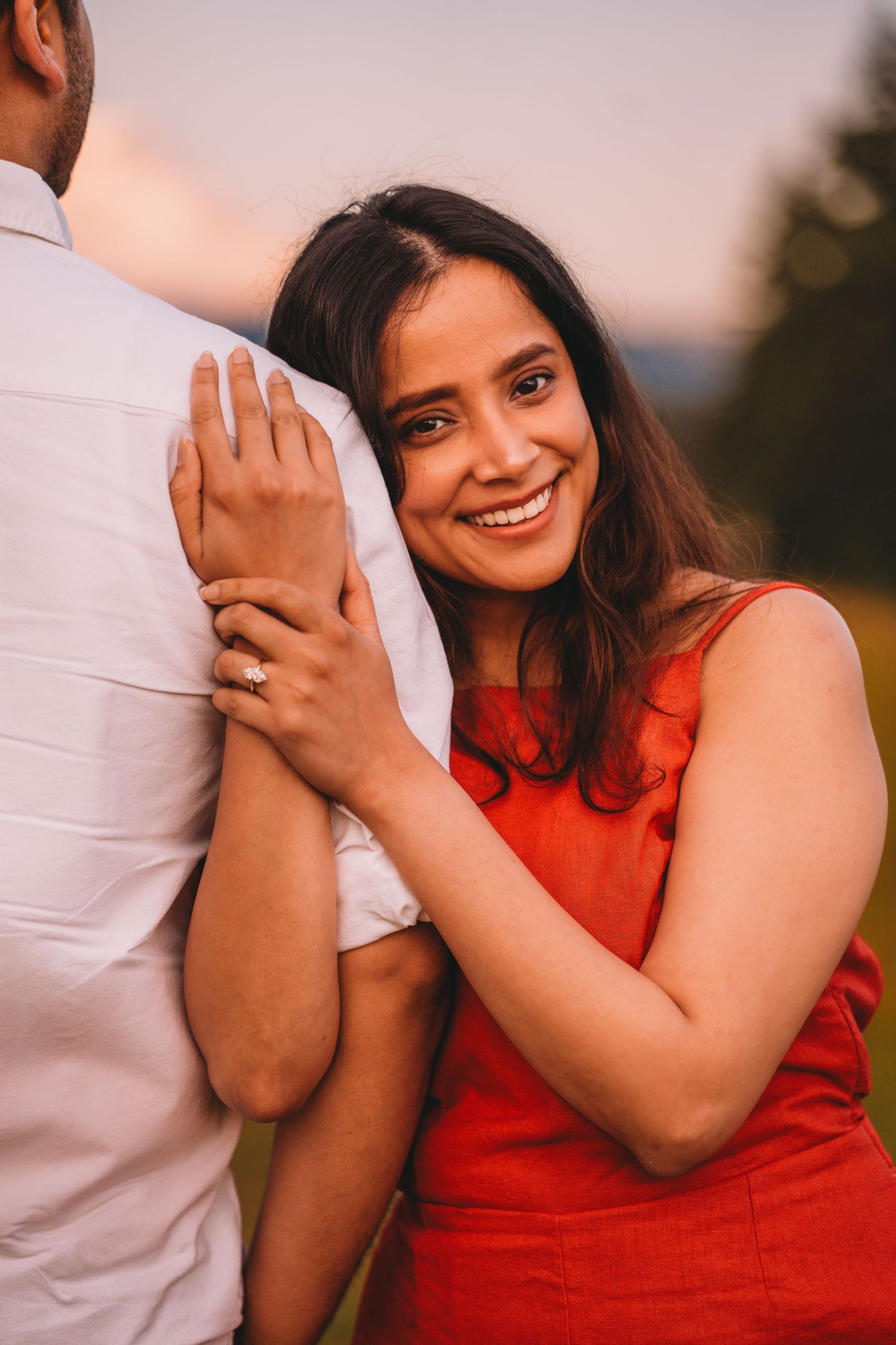 Smiling woman in red dress hugging man outdoors at sunset, wearing engagement ring.