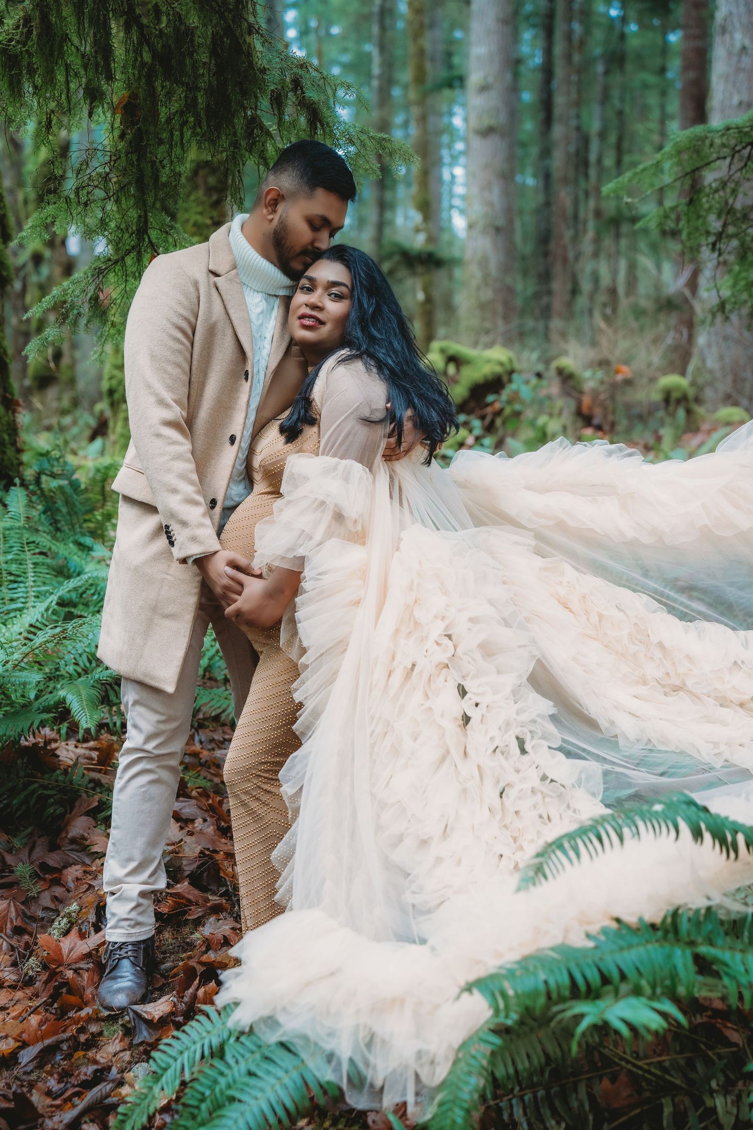 Couple in forest, woman in flowing ruffle gown, man in beige suit, embracing among ferns and trees.