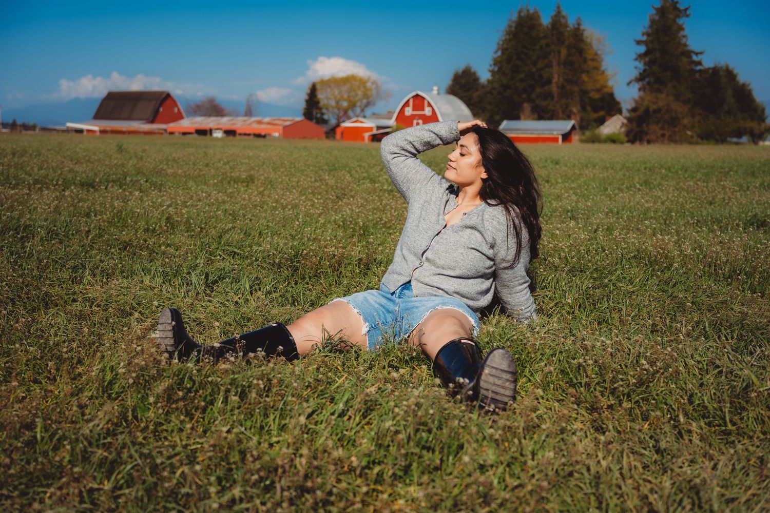 Woman in denim shorts and boots sitting in a green field with red farm buildings in the background on a sunny day.