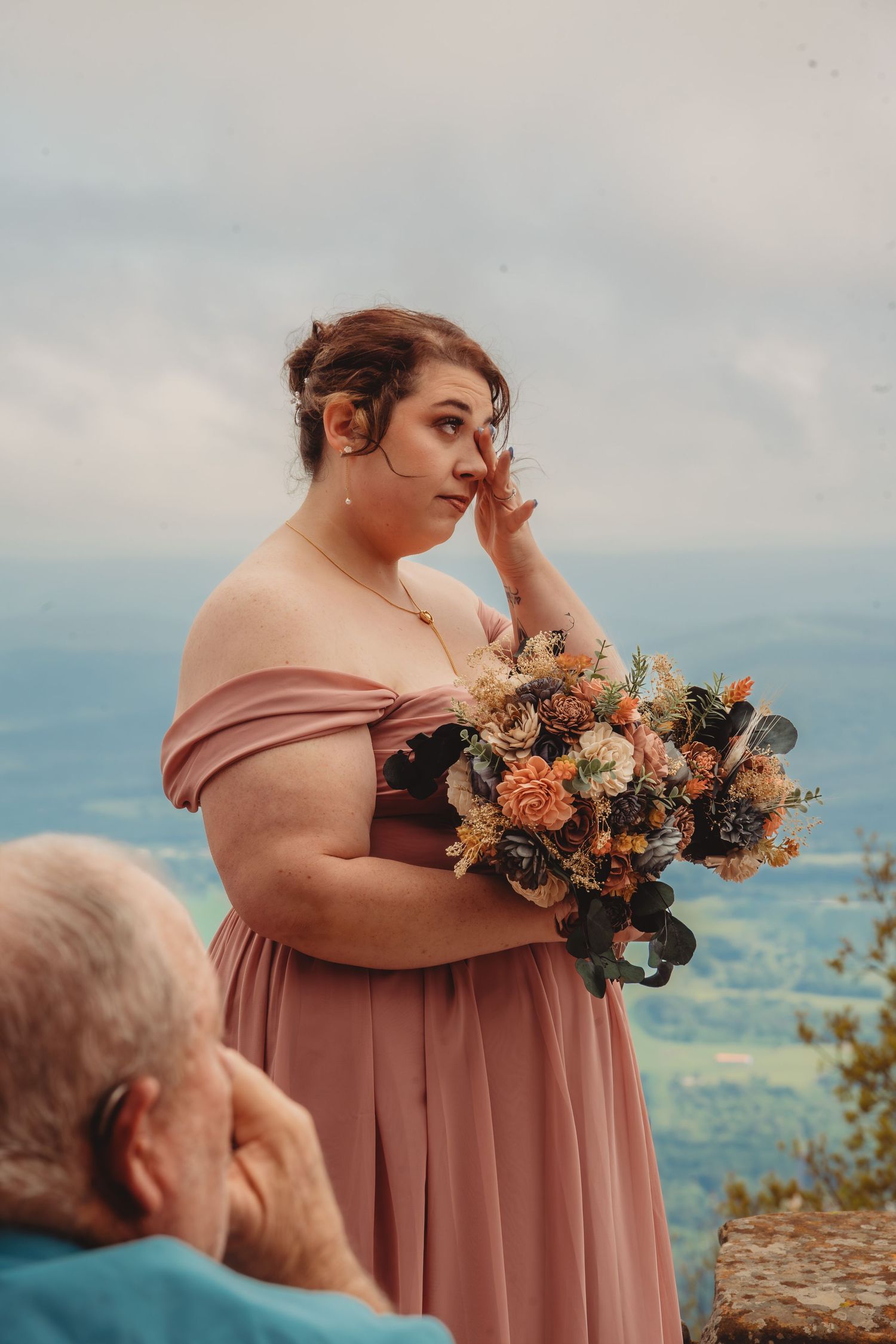 Woman in pink off-shoulder gown holding floral bouquet at scenic overlook, wiping tear at outdoor wedding.