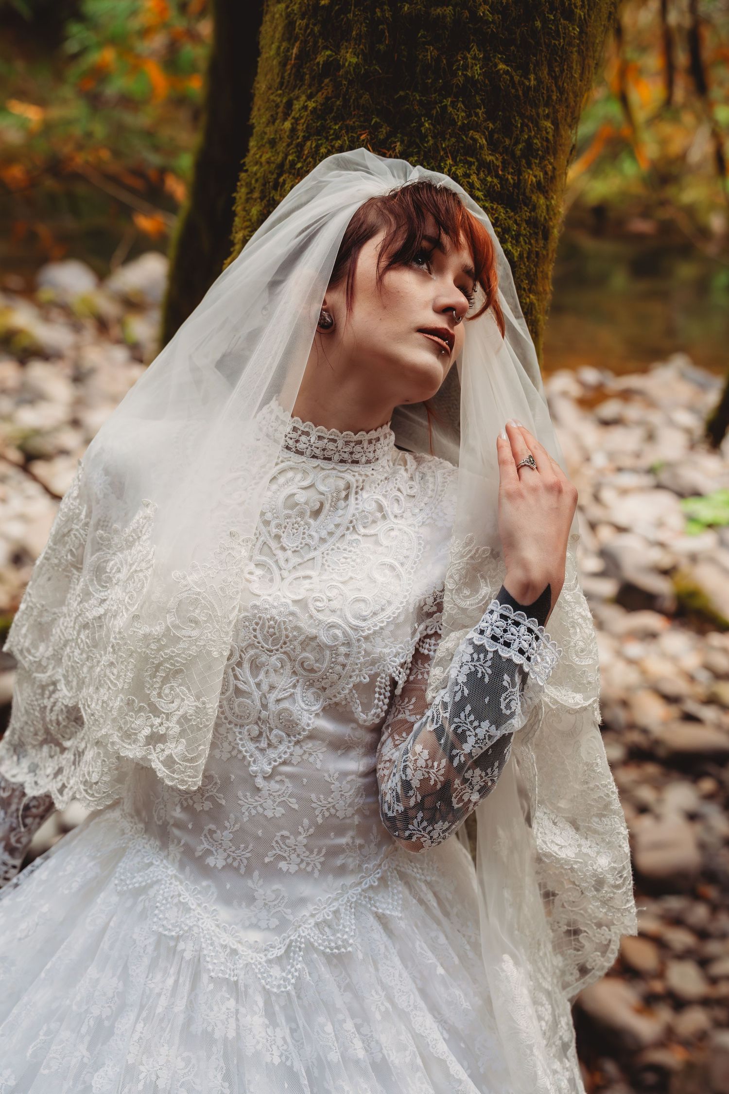 A bride in a vintage white lace gown and veil poses gracefully in an autumn forest setting near a mossy tree.