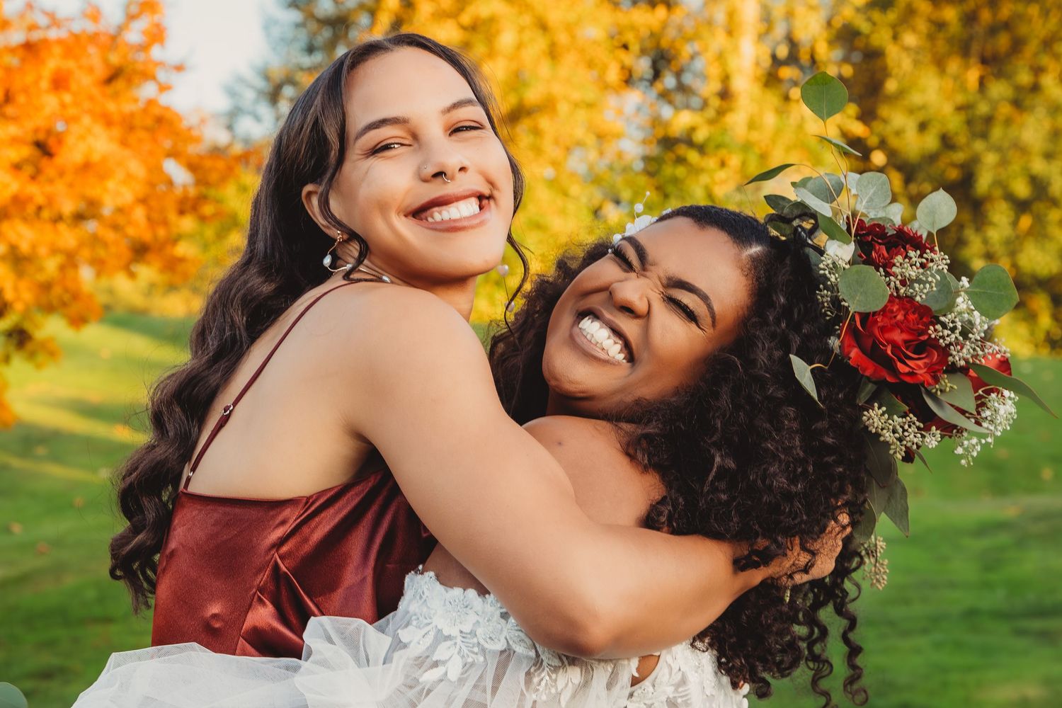 Two joyful women embrace outdoors in autumn, one wearing a floral crown and white dress, smiling brightly.
