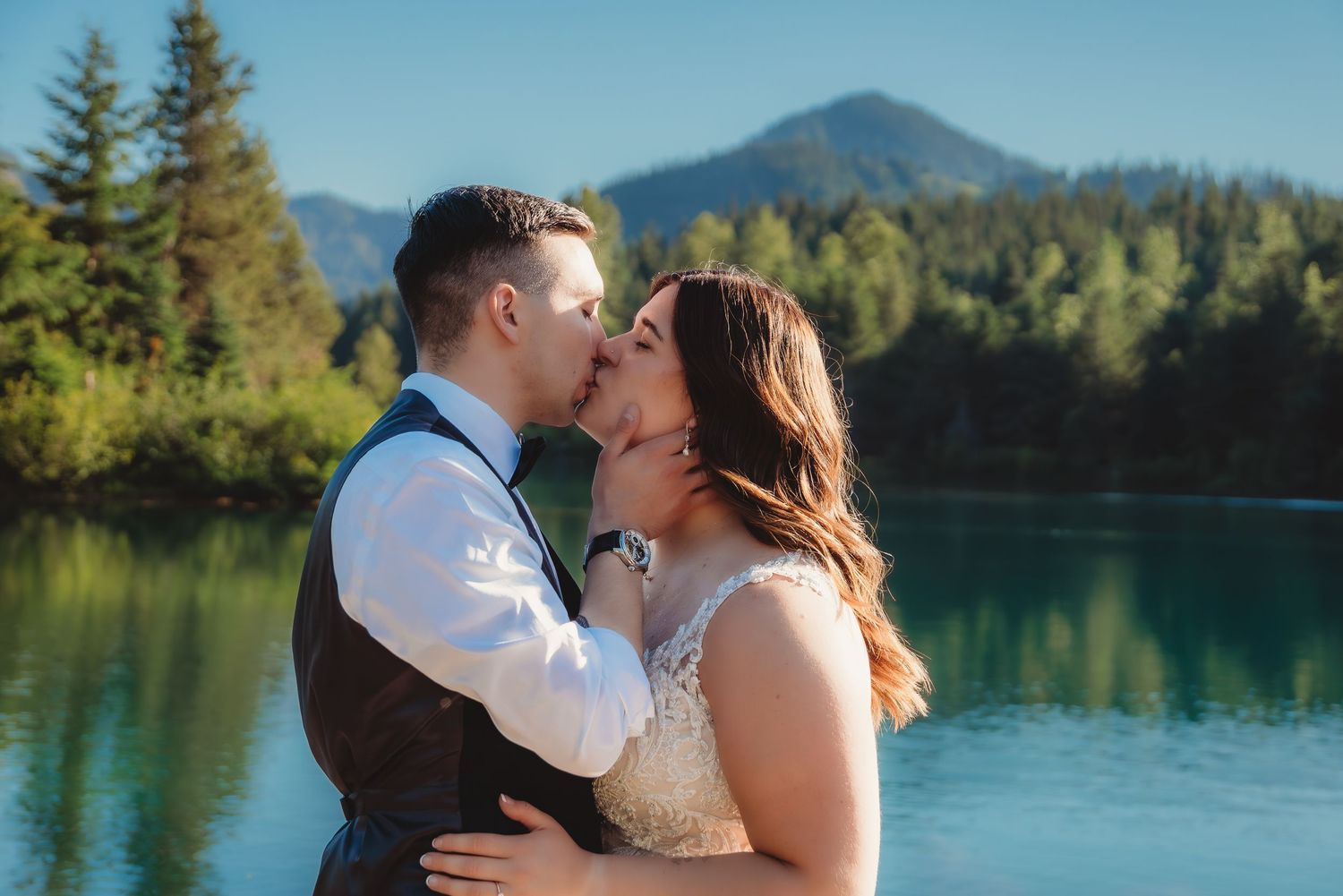 Bride and groom sharing a romantic kiss by a turquoise mountain lake surrounded by lush forest. gold creek pond