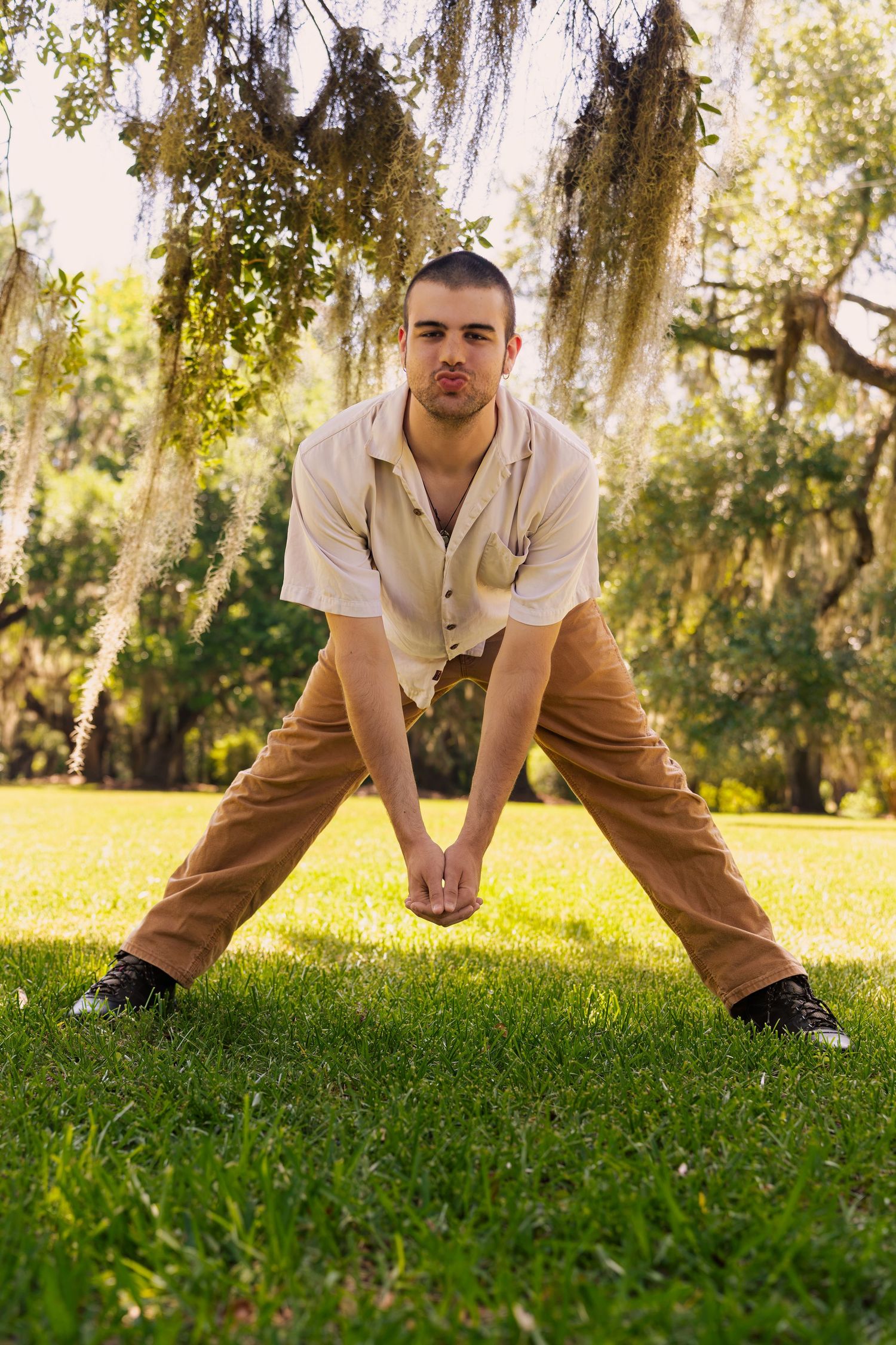 Young man in beige shirt and tan pants stretching on green grass under Spanish moss-draped trees in a sunny park.