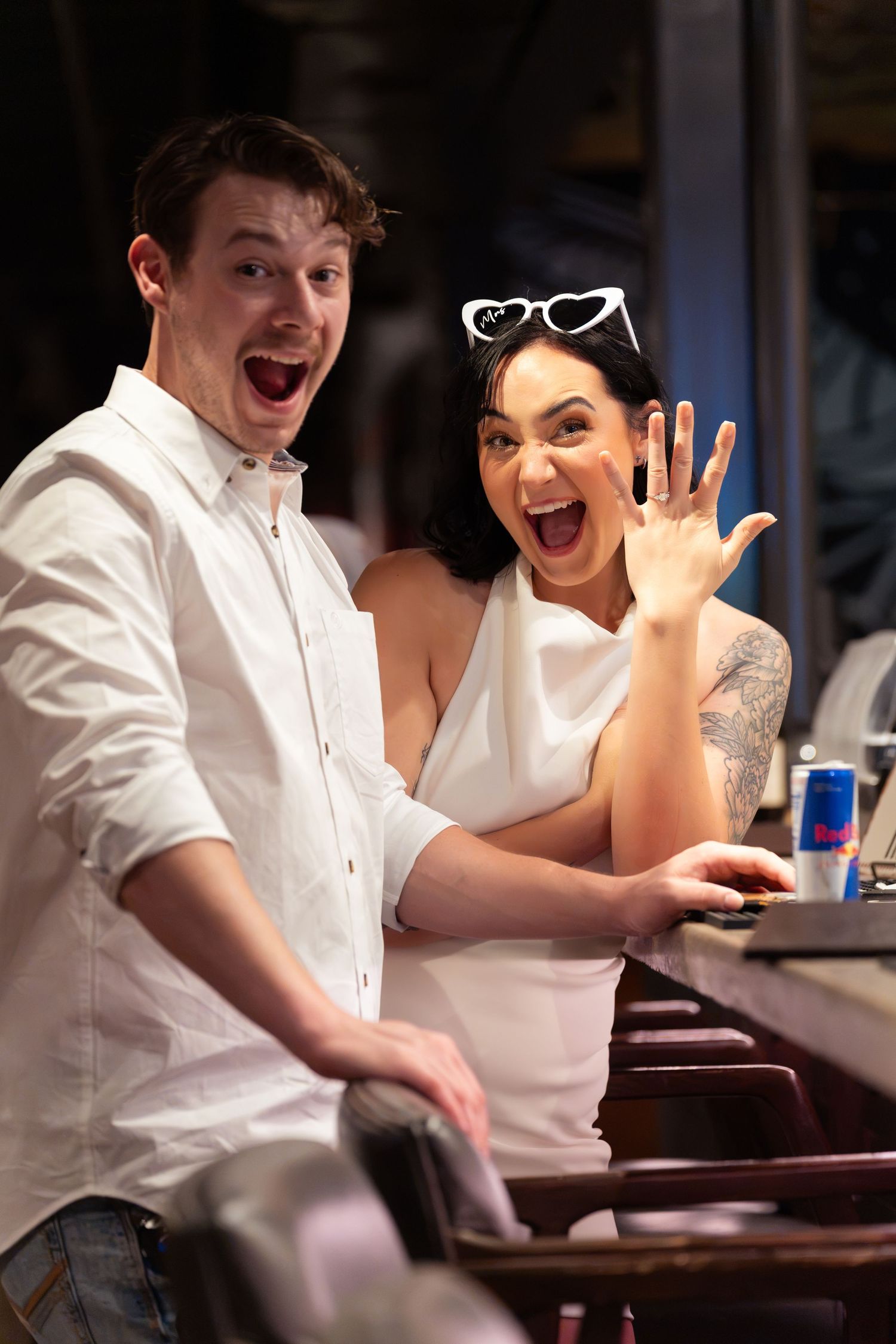 Excited couple at a bar, the woman showing her hand with a ring as both laugh, with heart sunglasses on her head.