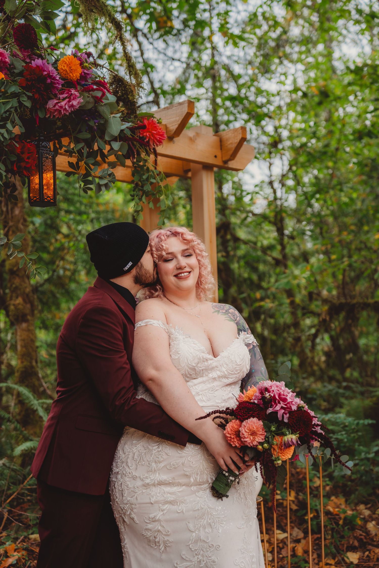 Bride in lace gown holds pink bouquet as groom in burgundy suit kisses her cheek under floral arch in forest.