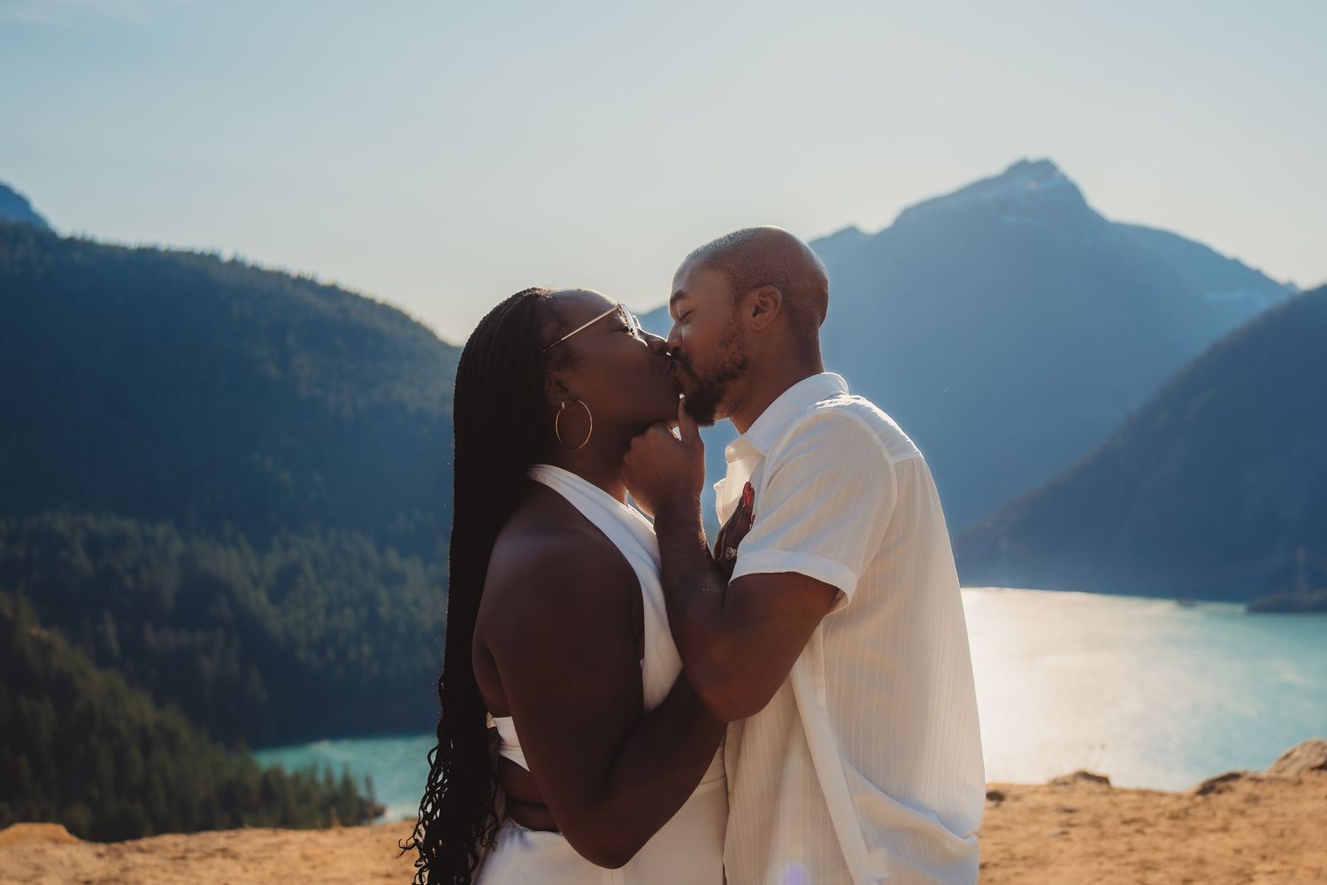 A couple shares a romantic kiss with a scenic mountain lake backdrop on a sunny day.