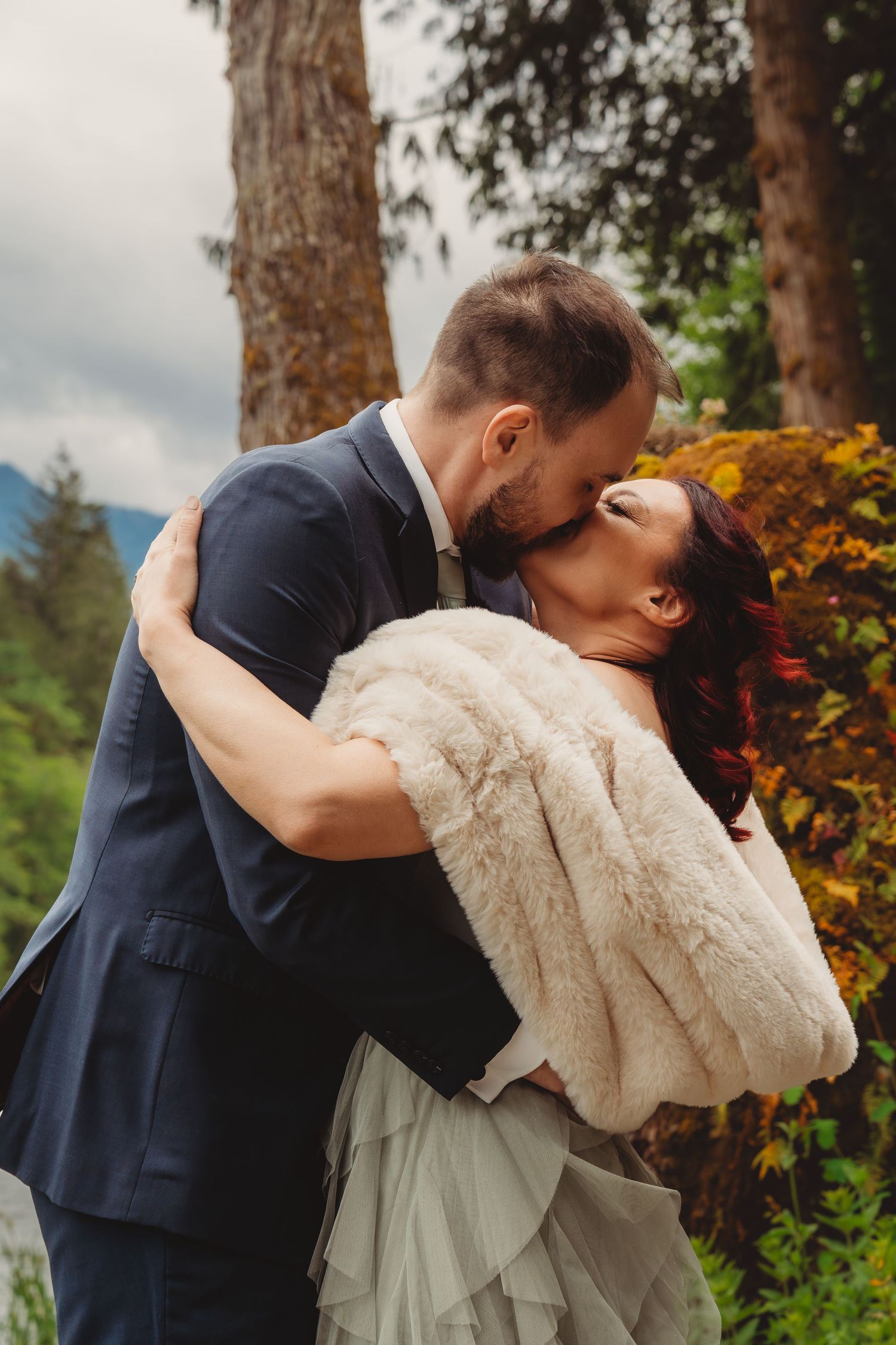 A bride and groom share a romantic kiss outdoors, surrounded by autumn trees and mountains, she wears a fur wrap.