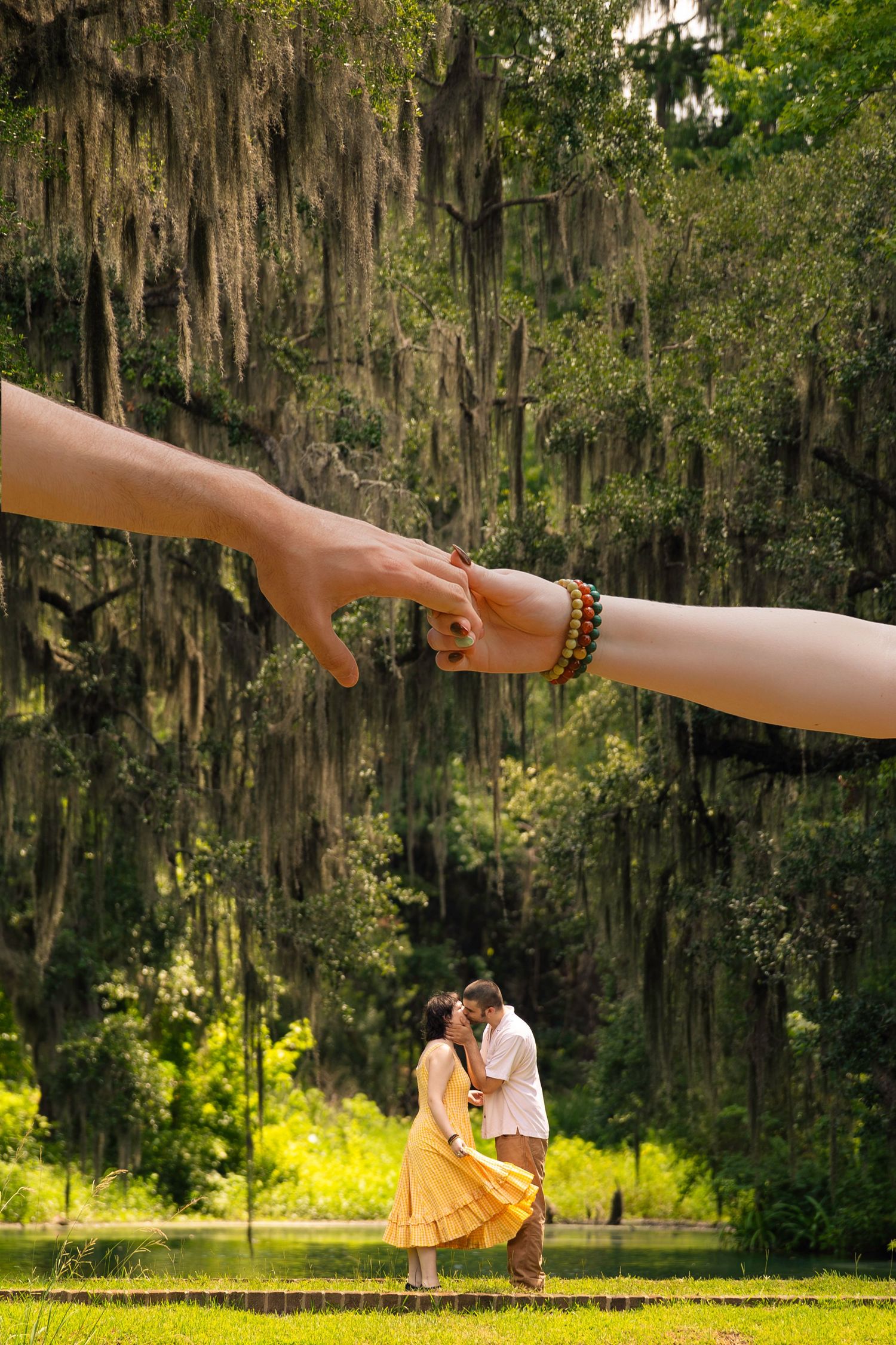 Couple kissing outdoors under moss-draped trees, with giant hands touching above them in a creative composite photo.