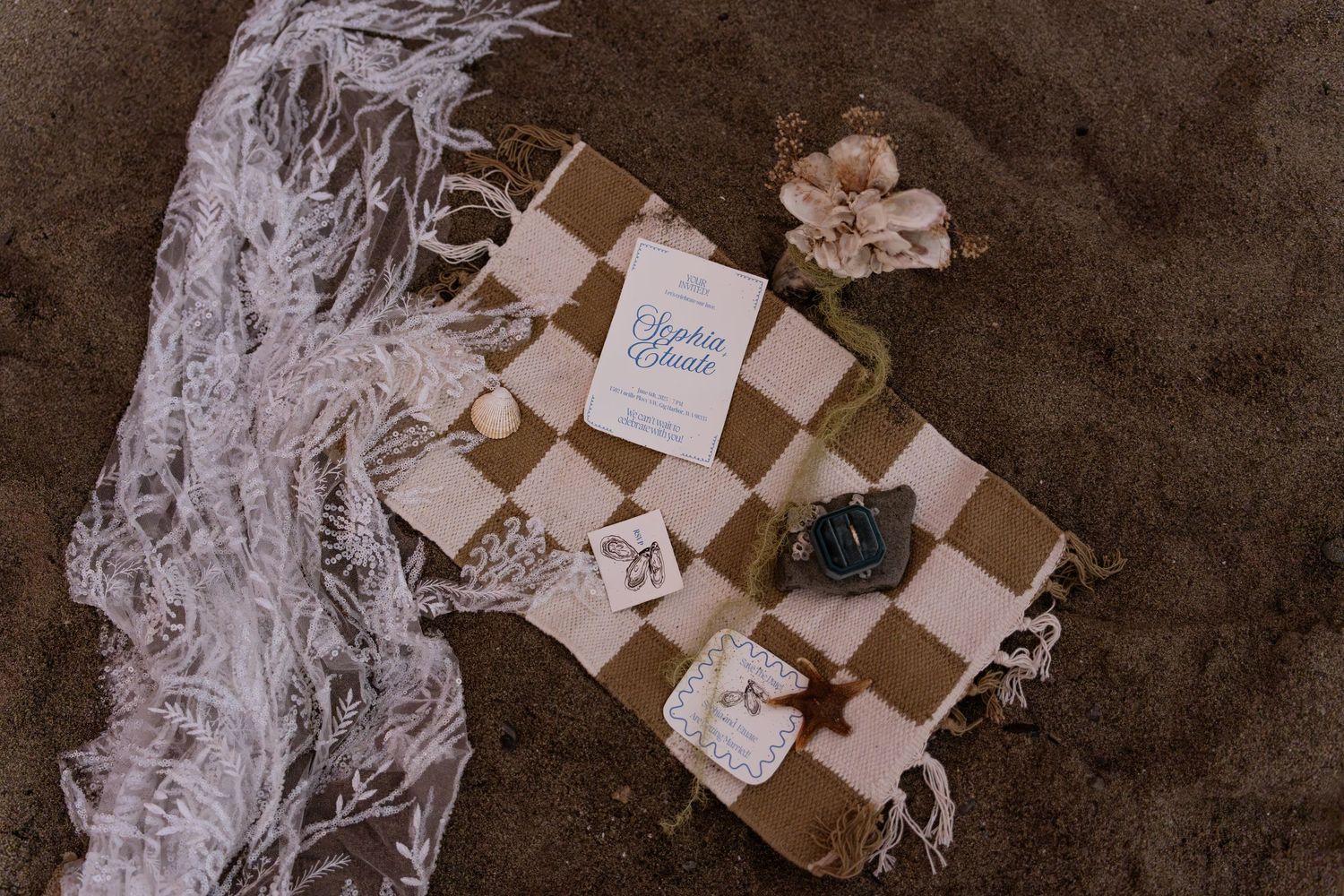 Checkered picnic blanket with wedding stationery, camera, and shells arranged on sandy beach.