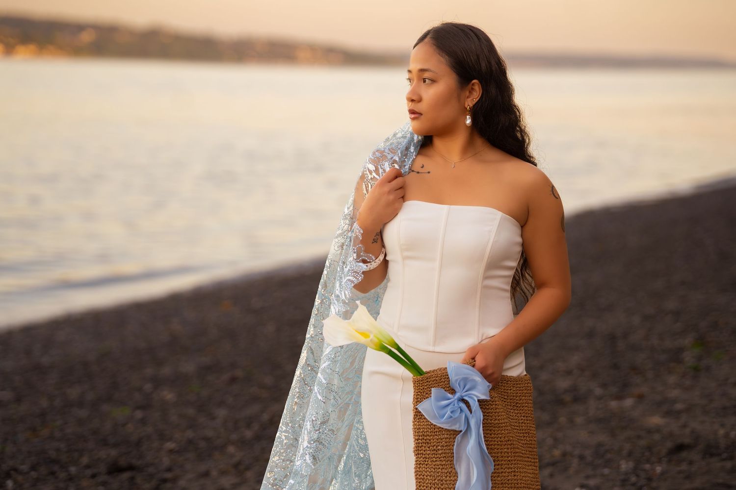 A woman in a white strapless gown holds a calla lily on a black sand beach at sunset.