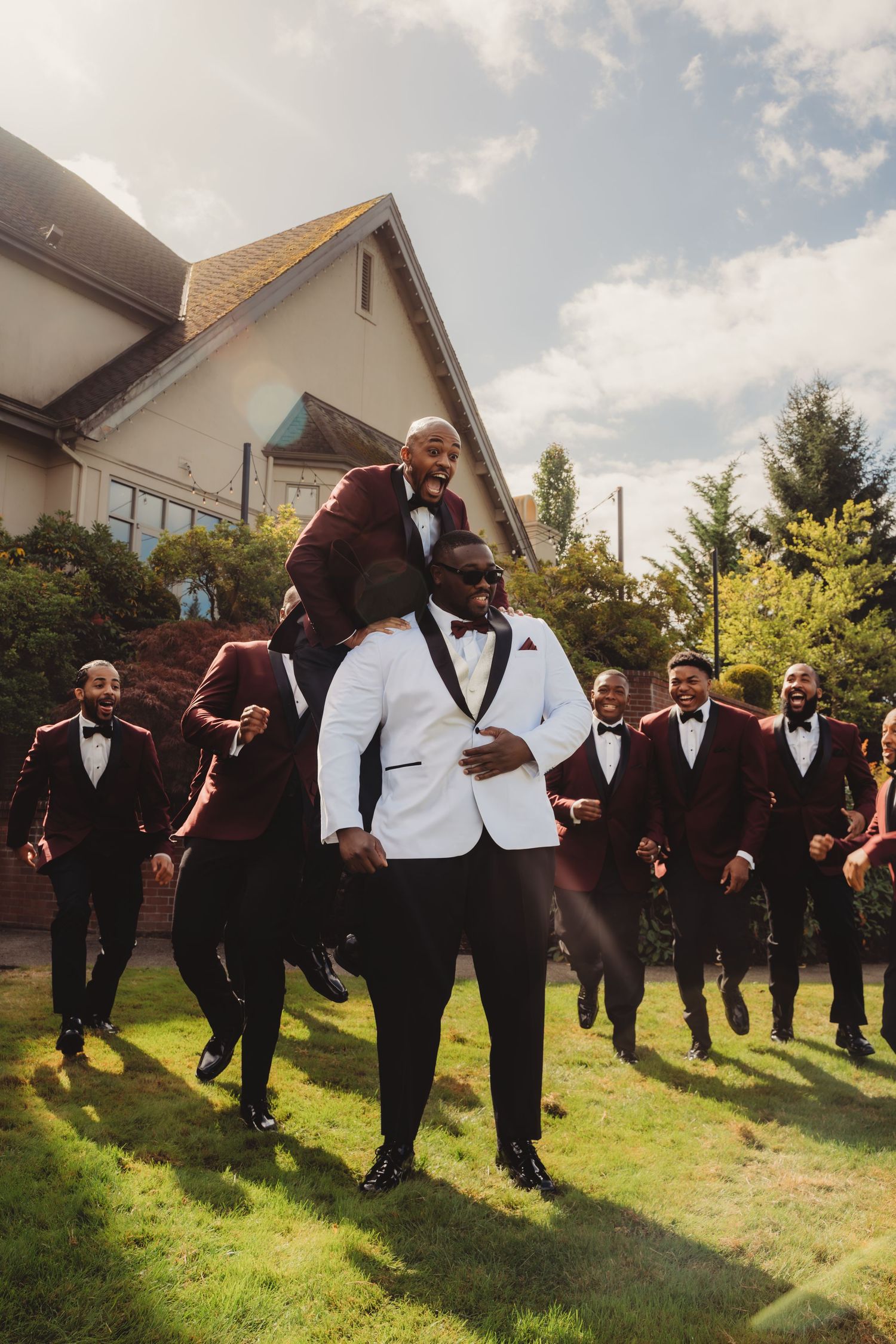 Groom in white tuxedo celebrates with groomsmen in burgundy suits outdoors on a sunny wedding day.