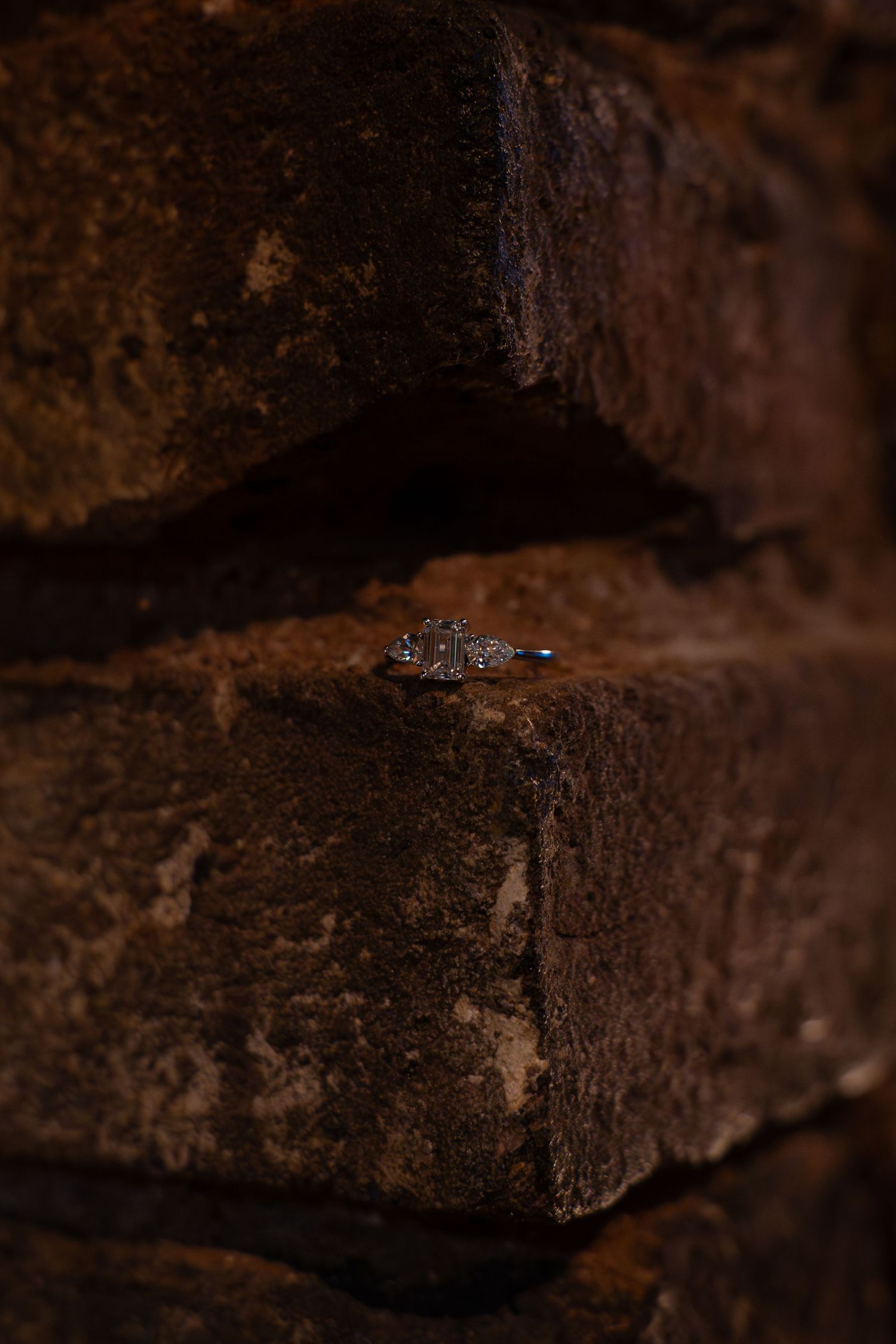 Sparkling diamond engagement ring resting on the ledge of a rustic, weathered brick wall in warm, moody lighting.