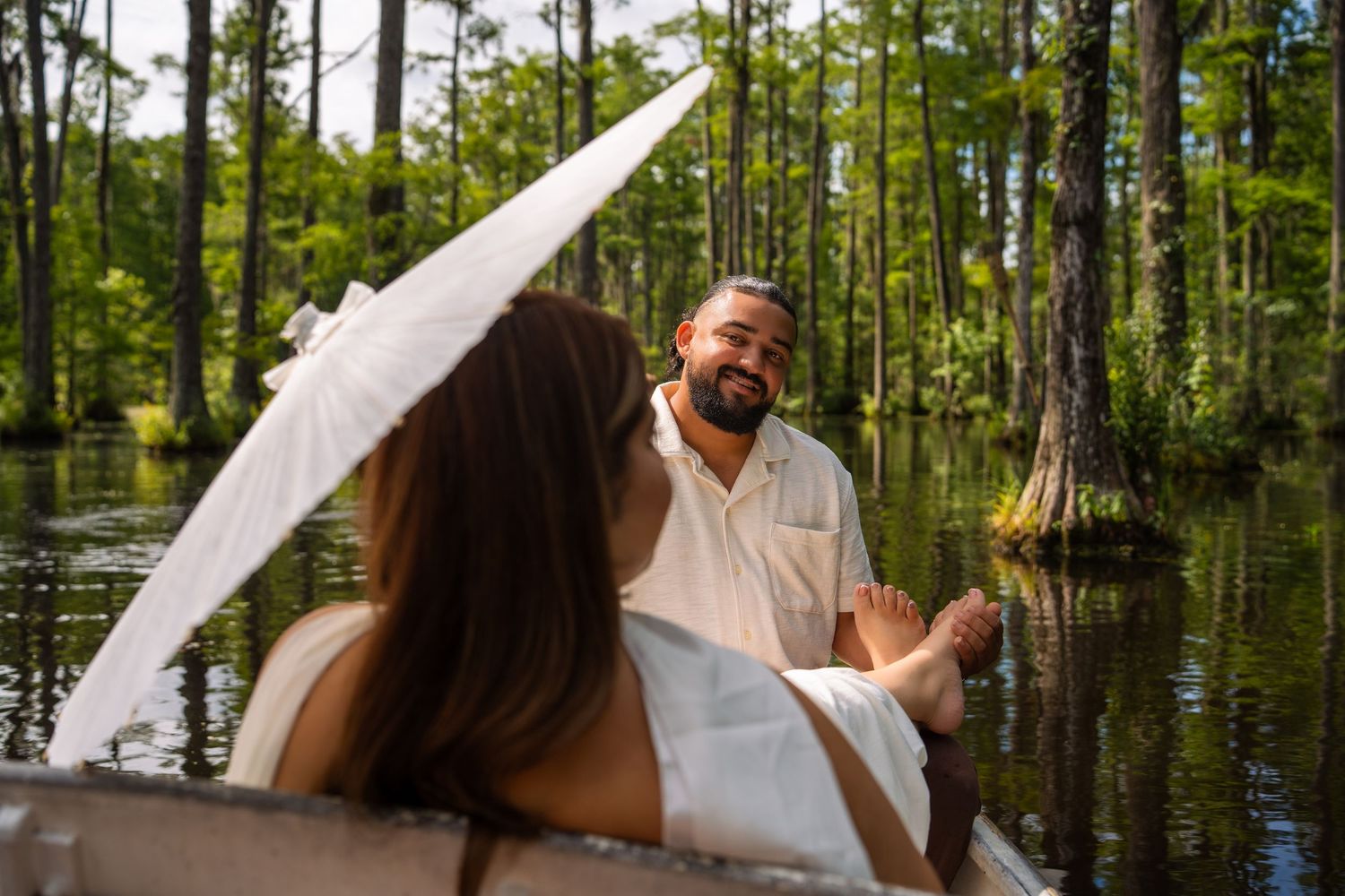 Couple in white attire sharing a romantic boat ride through a lush cypress swamp, smiling and holding hands.