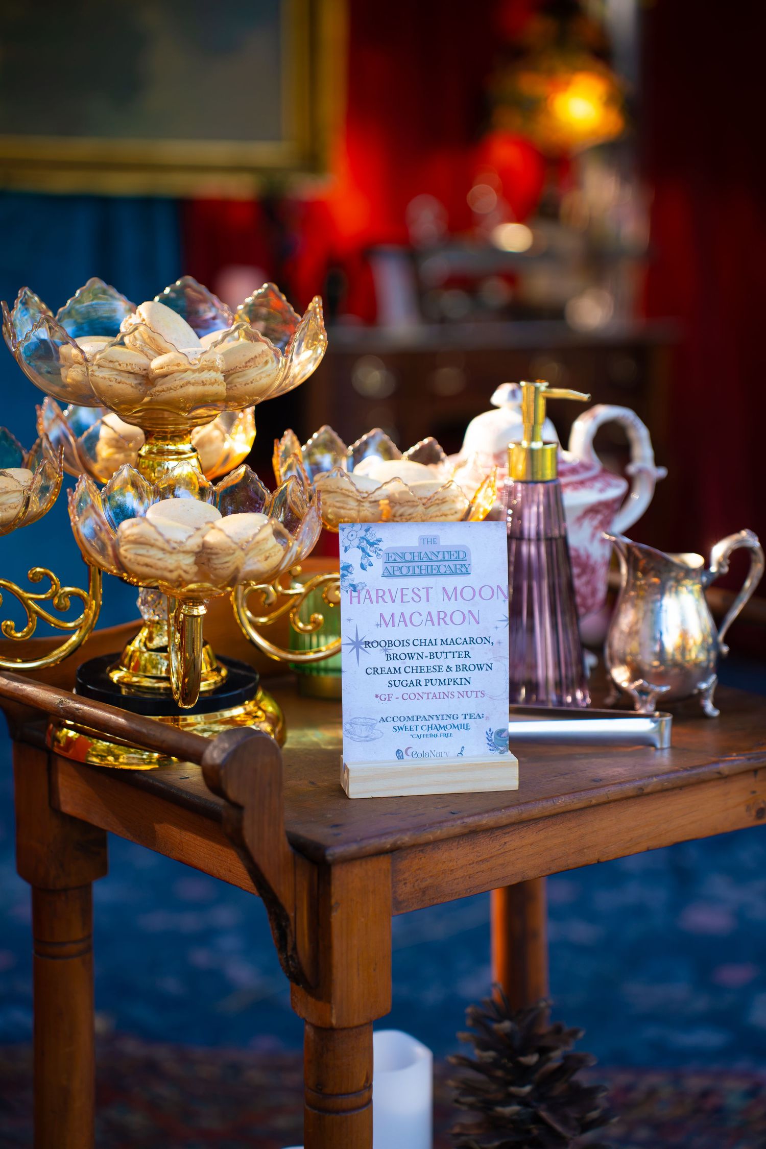 Gold tiered stand of macarons beside a Harvest Moon Macaron sign, a pink perfume bottle and a pitcher on a wooden table.