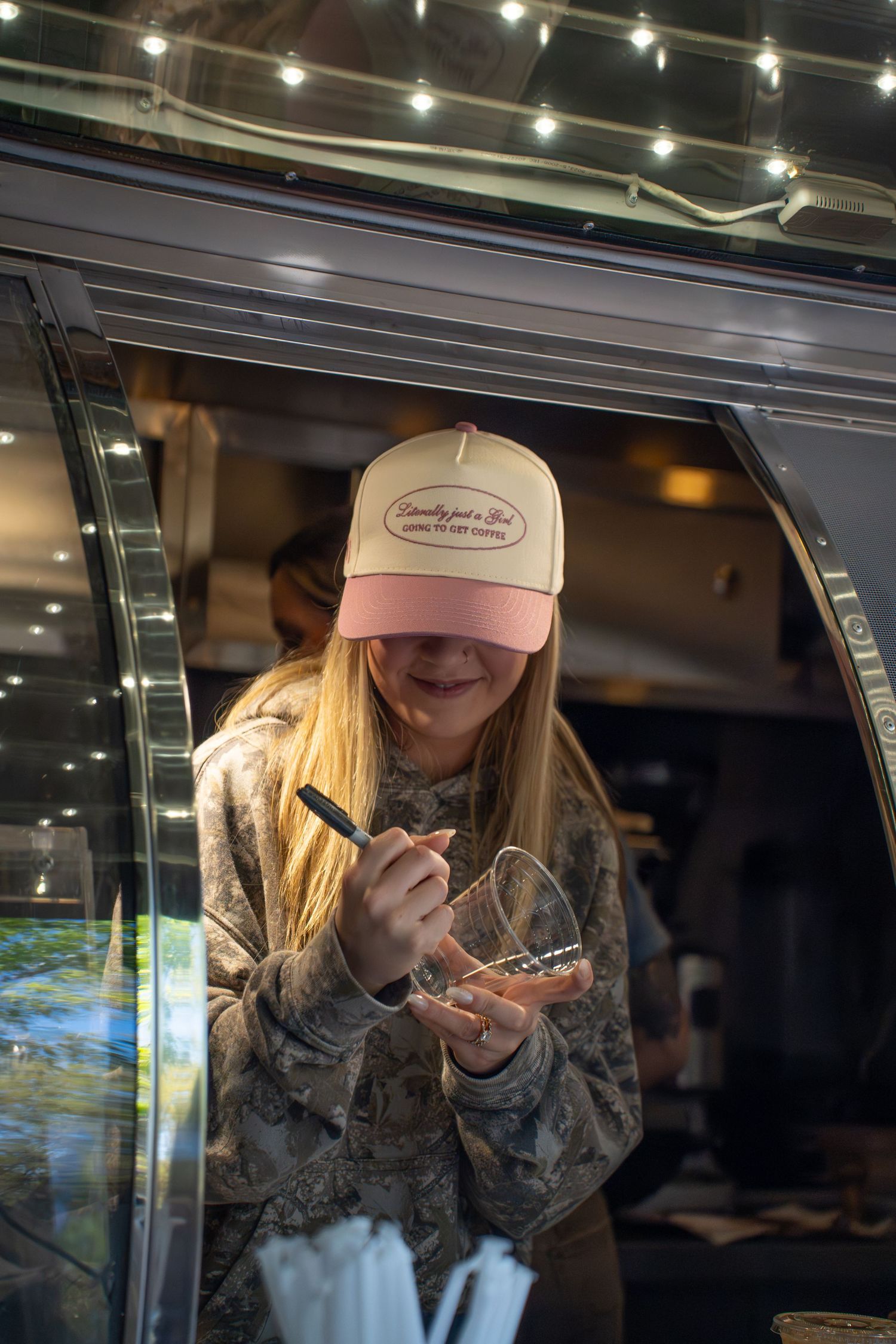 Smiling woman in pink trucker hat and camo hoodie writing on glass at a food truck with string lights.