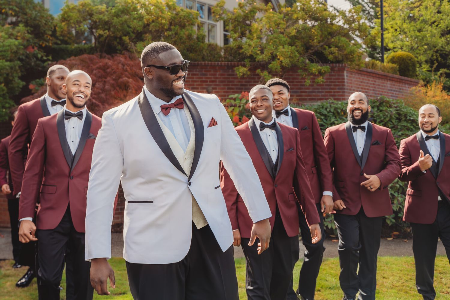 Groom in white tuxedo jacket leads smiling groomsmen in matching burgundy tuxedos outdoors at wedding celebration.