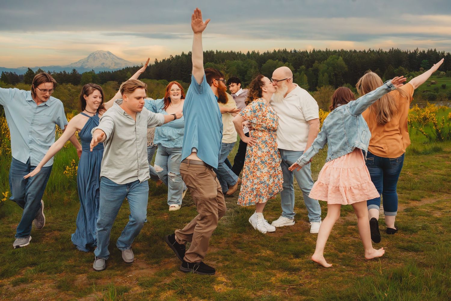 Large group of people dancing joyfully outdoors in a field with mountain and forest backdrop at sunset.