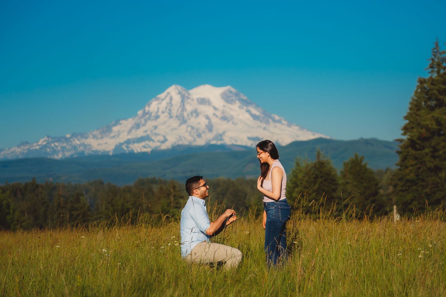 Man proposes to woman in a sunny meadow with snow-capped Mount Rainier in the background on a clear day.