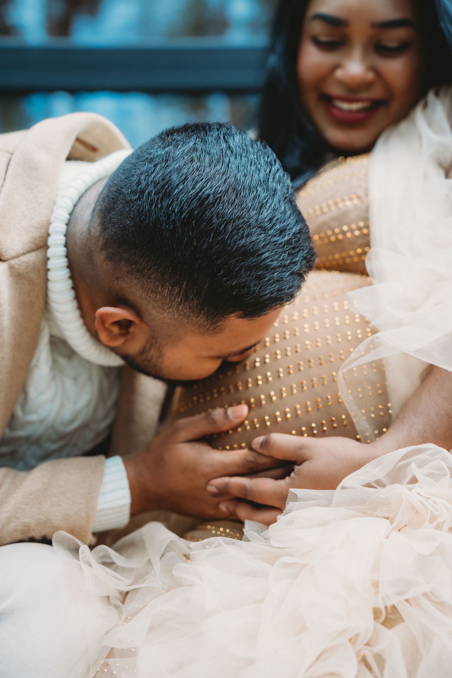 A man tenderly kisses a pregnant woman's baby bump while she smiles joyfully during a maternity photoshoot.
