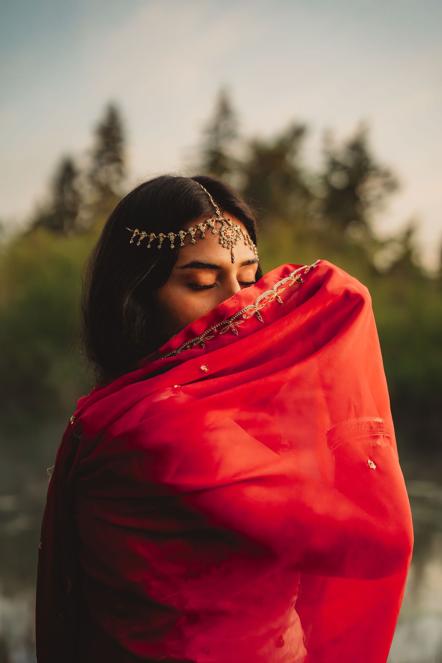 Woman in red saree with gold maang tikka jewelry, eyes closed, standing near water with trees in background.