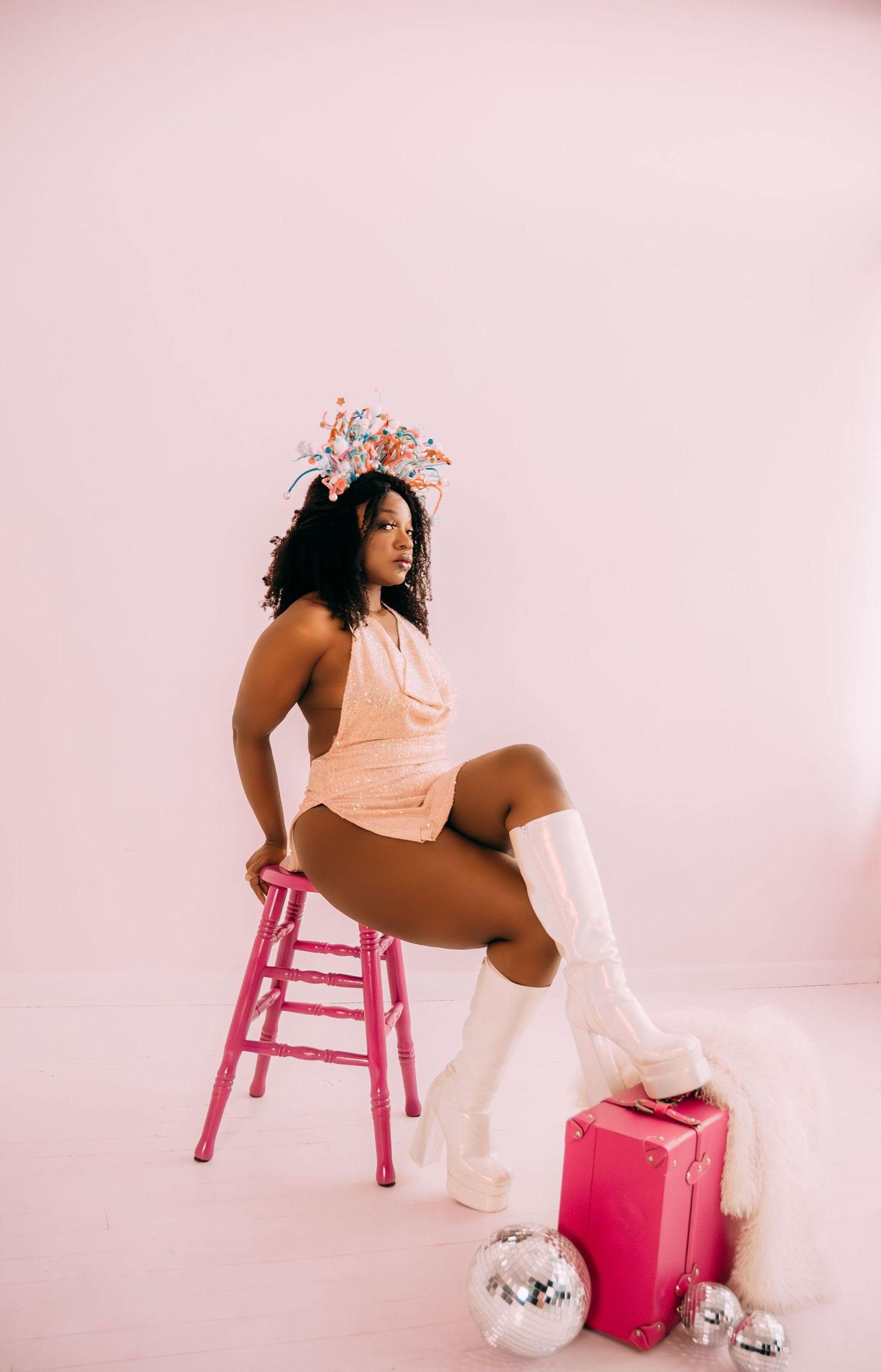 Woman in pink bodysuit sitting on pink stool with floral crown, white boots, and disco balls.