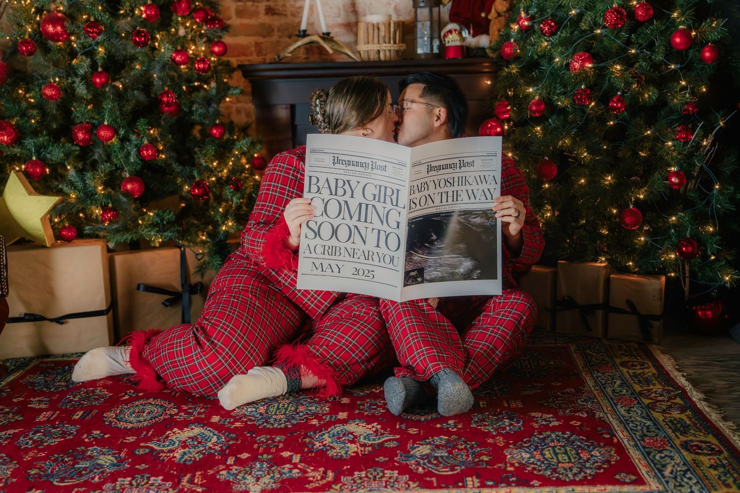 Couple in matching red plaid pajamas holding baby girl pregnancy announcement newspaper by Christmas tree.