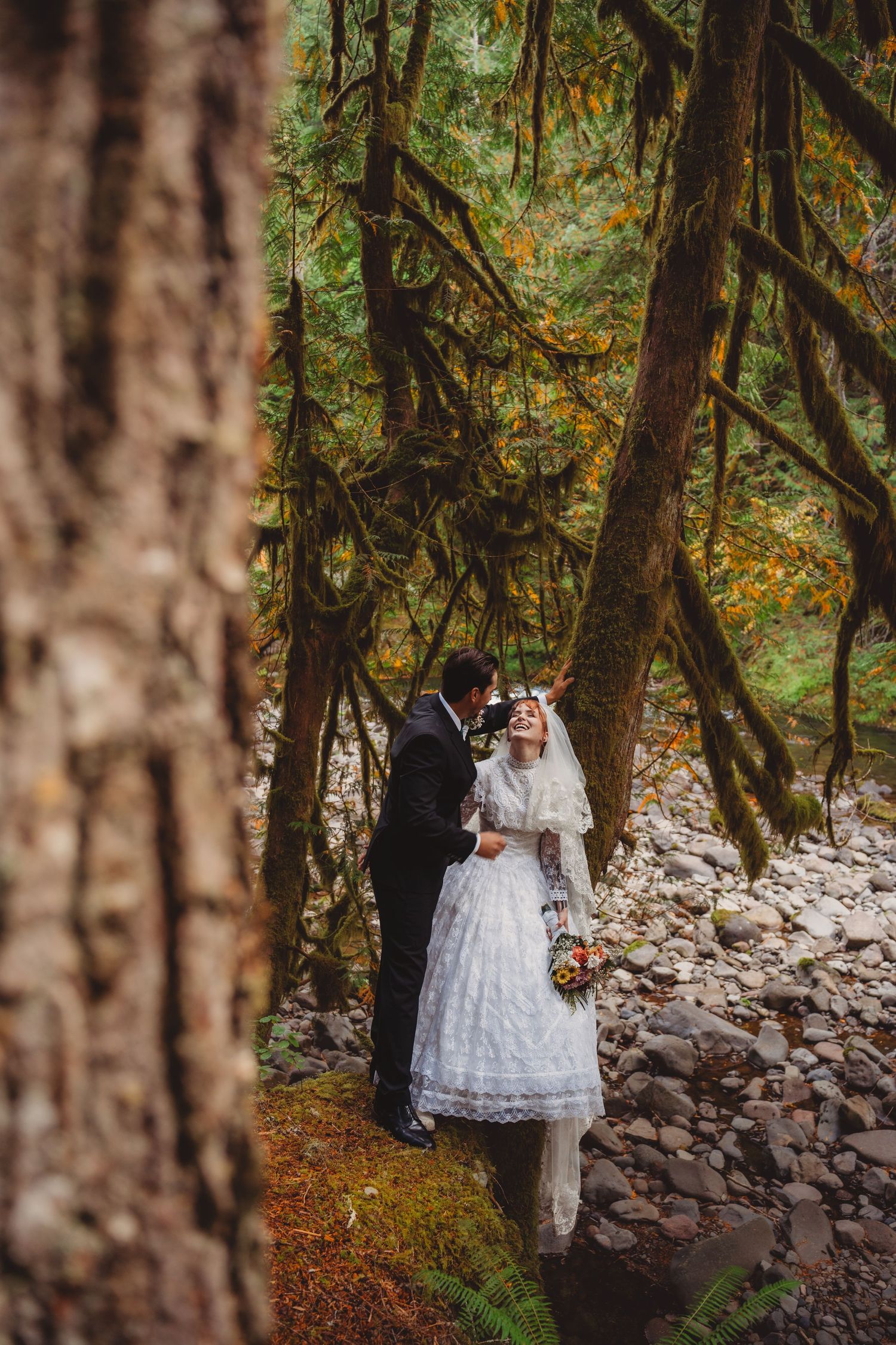 Bride and groom share a romantic moment in a lush mossy forest, bride wearing a vintage lace dress and floral crown.