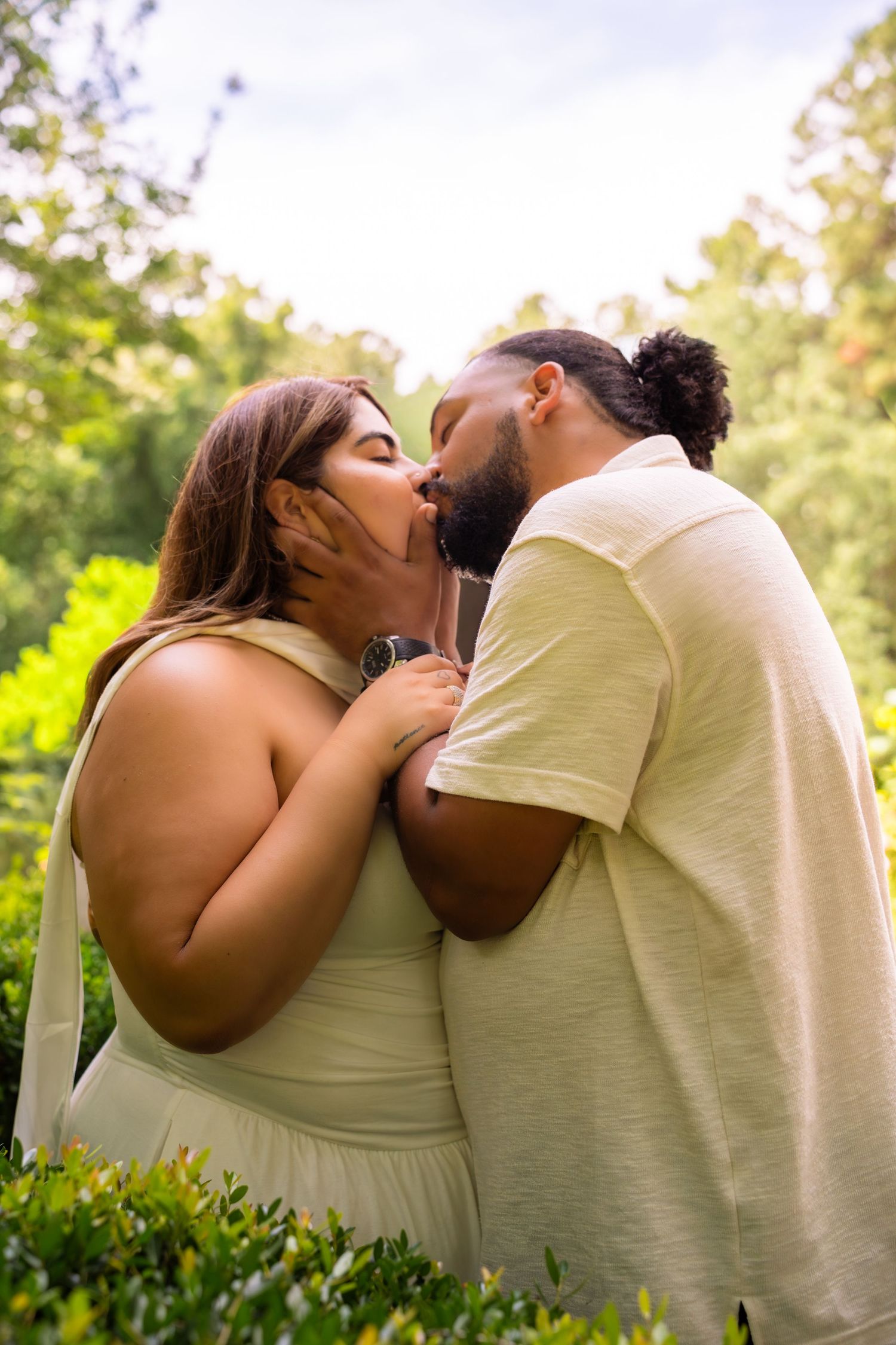A couple shares a romantic kiss outdoors surrounded by lush green trees and bushes on a warm sunny day.