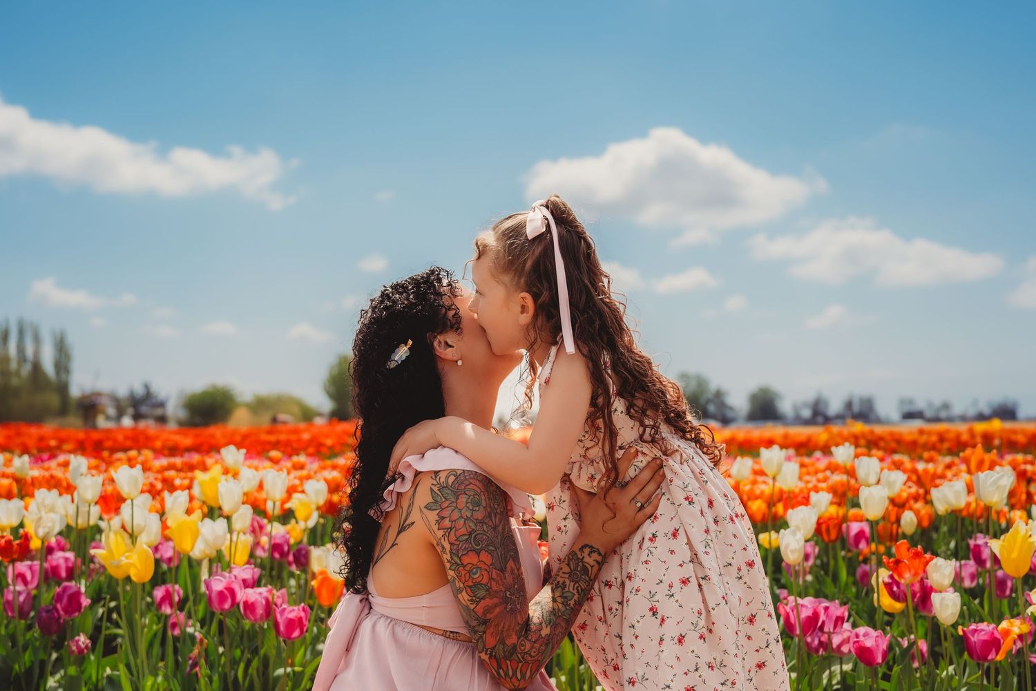 A tattooed mother kisses her young daughter in a vibrant tulip field under a blue sky.