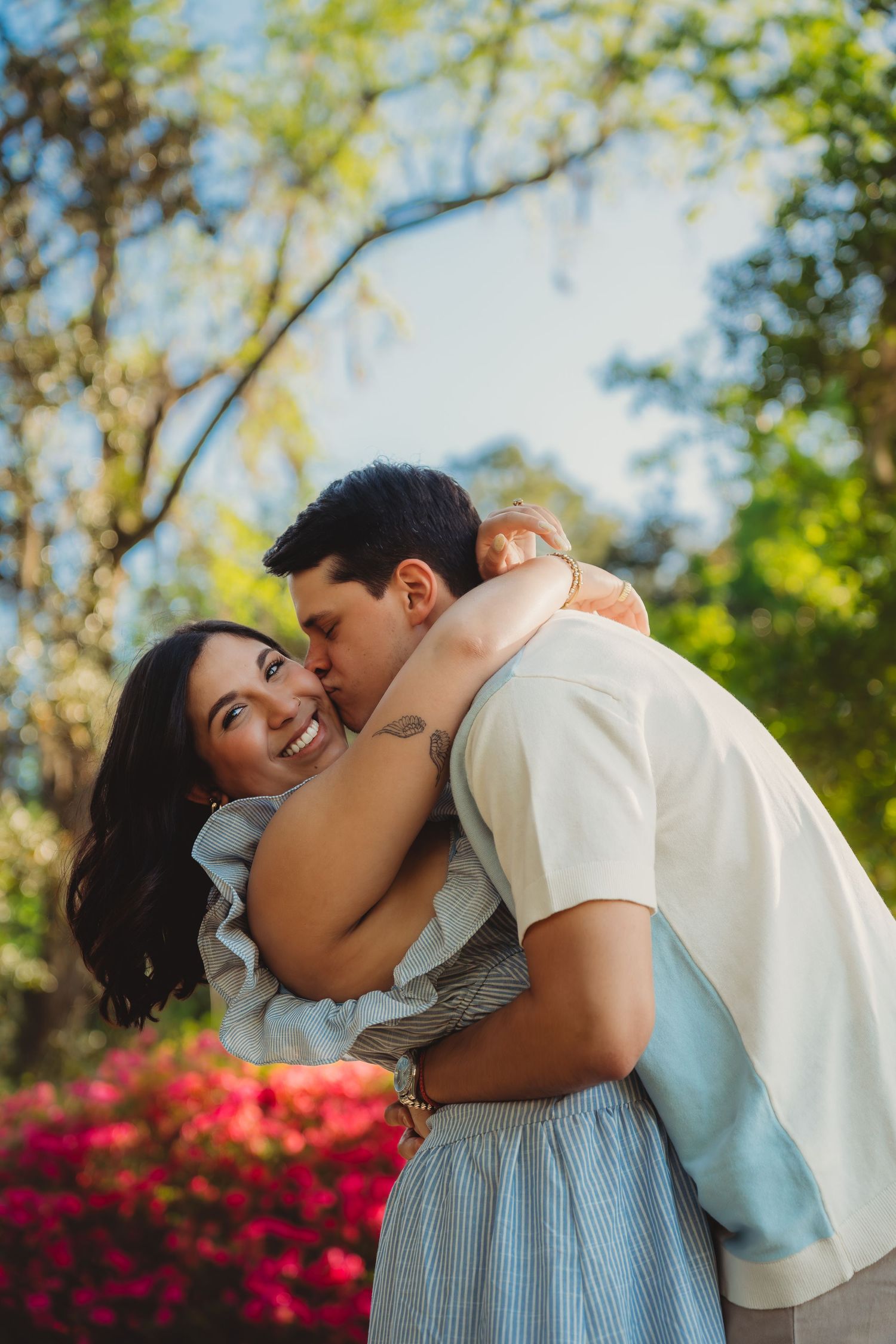 A couple embraces outdoors, woman smiling brightly as man kisses her cheek near vibrant red flowers and lush green trees.