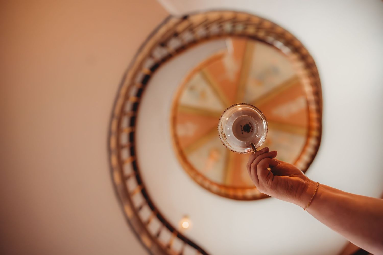Hand holding a vintage magnifying glass beneath a spiral staircase with ornate wooden railings viewed from below.