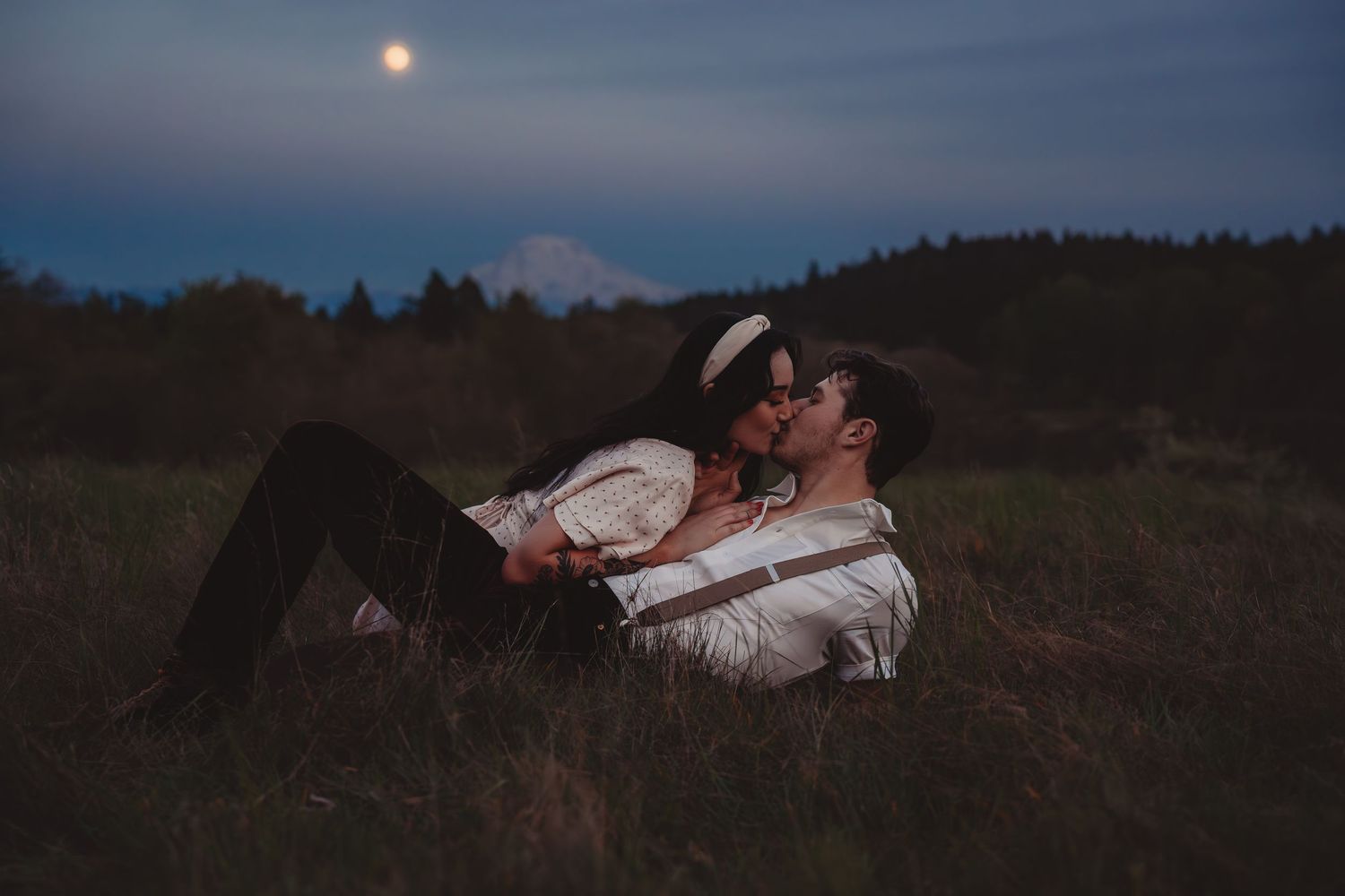 Couple kissing romantically in a moonlit field at dusk with misty mountains and trees in the background.